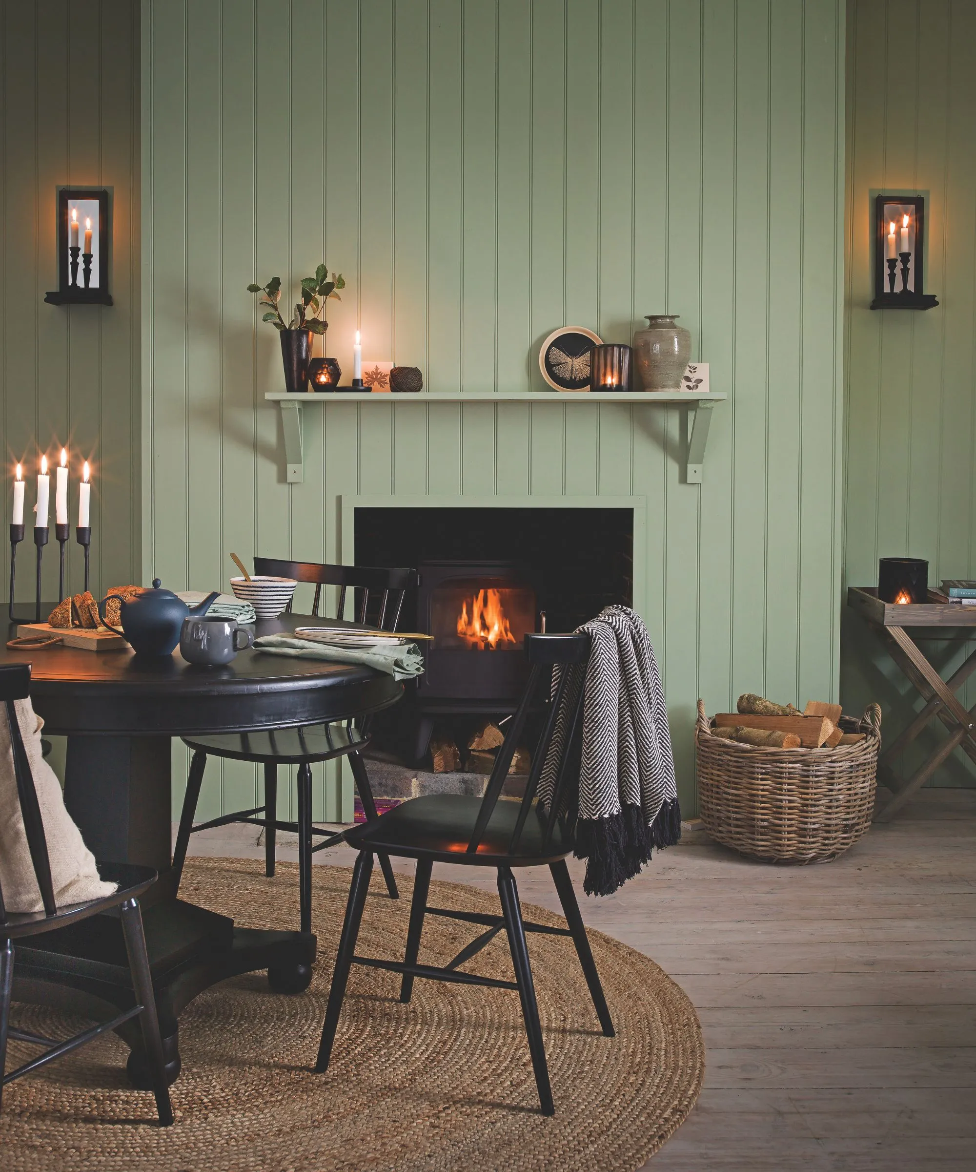 Salon wyłożony zielonymi panelami z płonącym w kominku rattanowym okrągłym dywanikiem, okrągłym czarnym stołem jadalnym z dopasowanymi krzesłami, wiklinowym koszem drewna opałowego i zapalonymi świecami stożkowymi.' loading='lazy' title=