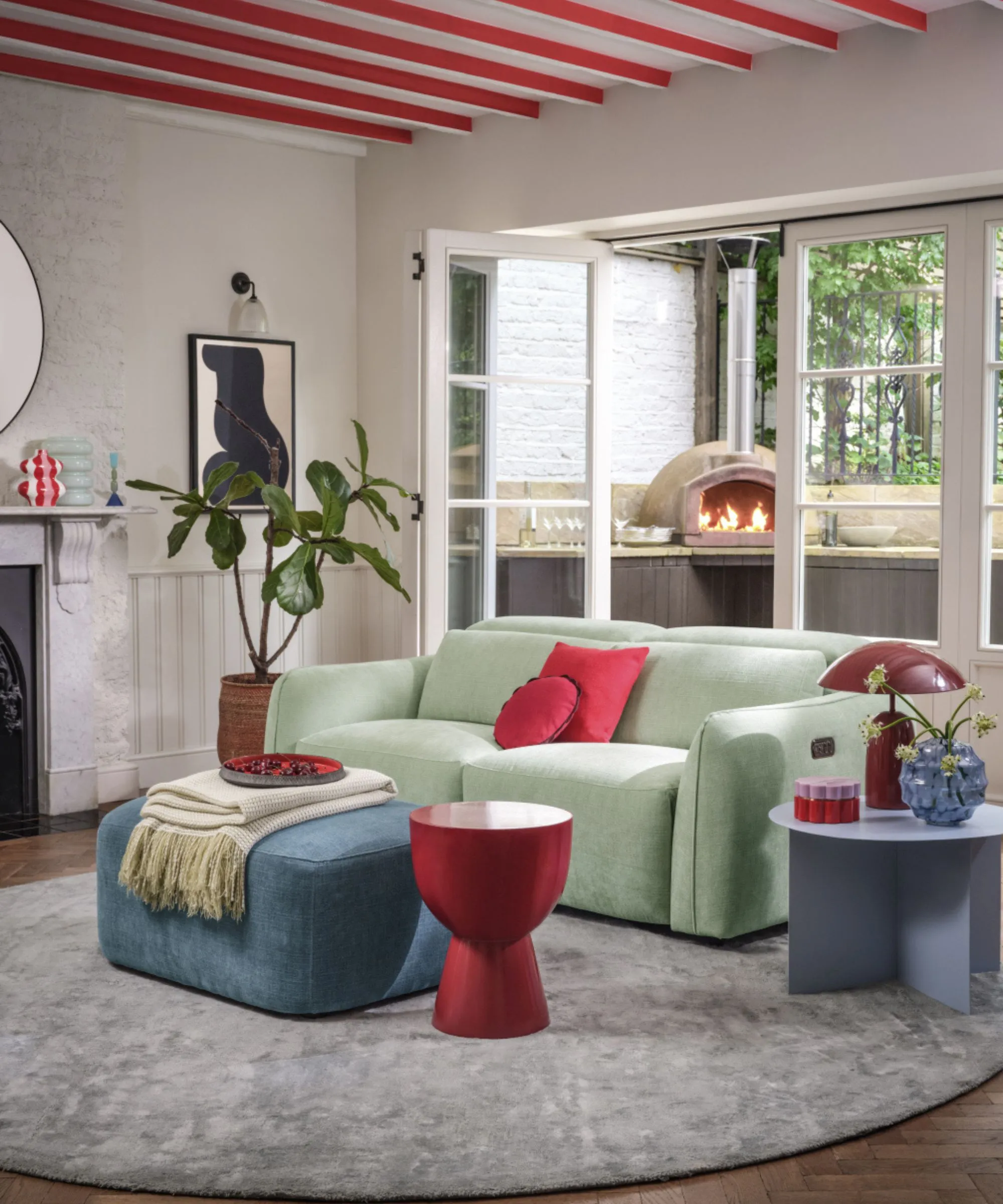 Lillehytte -stue med røde malede loftsbjælker og matchende accenter inklusive puder sideborde Konisk lampe og mantelindretning. De franske døre er åbne i en lille gårdsplads med pizzaovn, der brænder' loading='lazy' title=