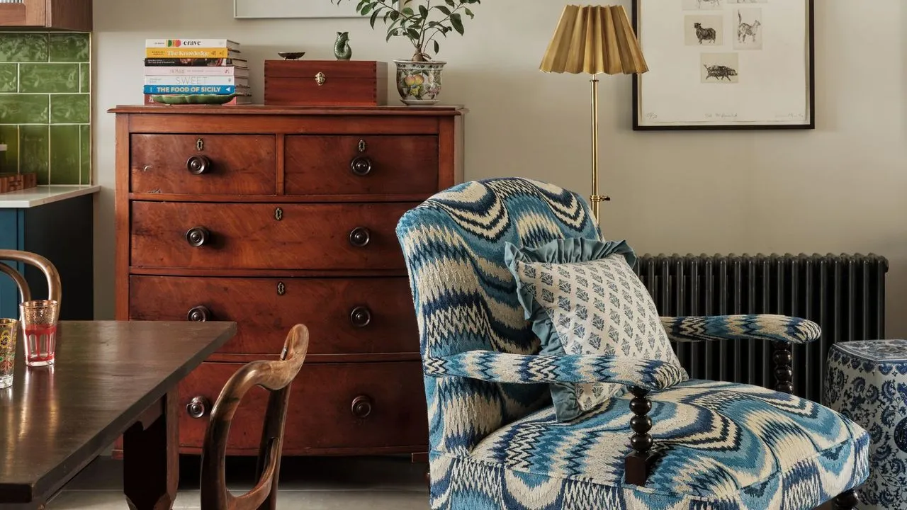 オープンプランのキッチンダイナー。キッチンキャビネットの一角に緑色の地下鉄のスプラッシュバックタイルが見えます。正面と中央には、ボビンウッドの特徴を備えたブルーとクリーム色の模様入りの布張りのアームチェアがあります。背景には黒いラジエーターと、ミッドセンチュリーの 5 段引き出しのキャビネットが見えます。' loading='lazy' title=