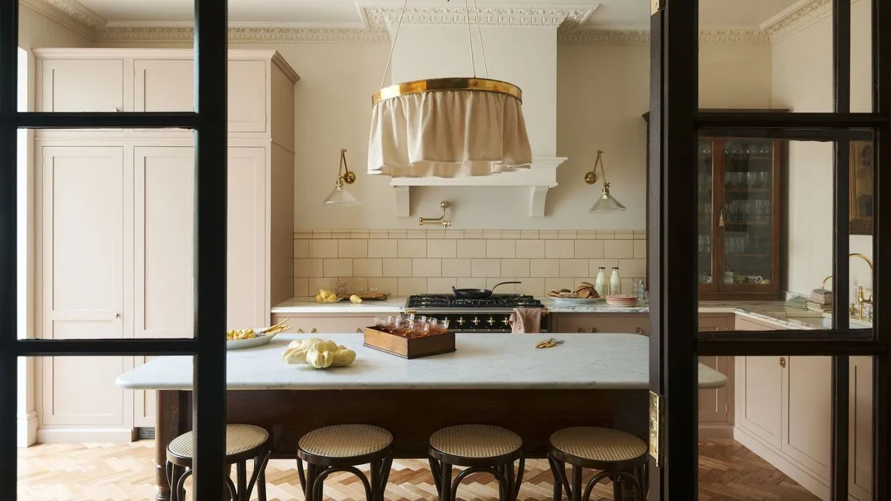 内部の黒い Crittal ドアを通して見た、ピーチピンクのキャラクターキッチン。大理石のアイランドテーブルと背もたれのない木製のバースツールが見えます。背景には、スプラッシュバックタイル張りのストーブエリアと、ピンクとガラスの前面茶色のキャビネットが組み込まれています。' loading='lazy' title=