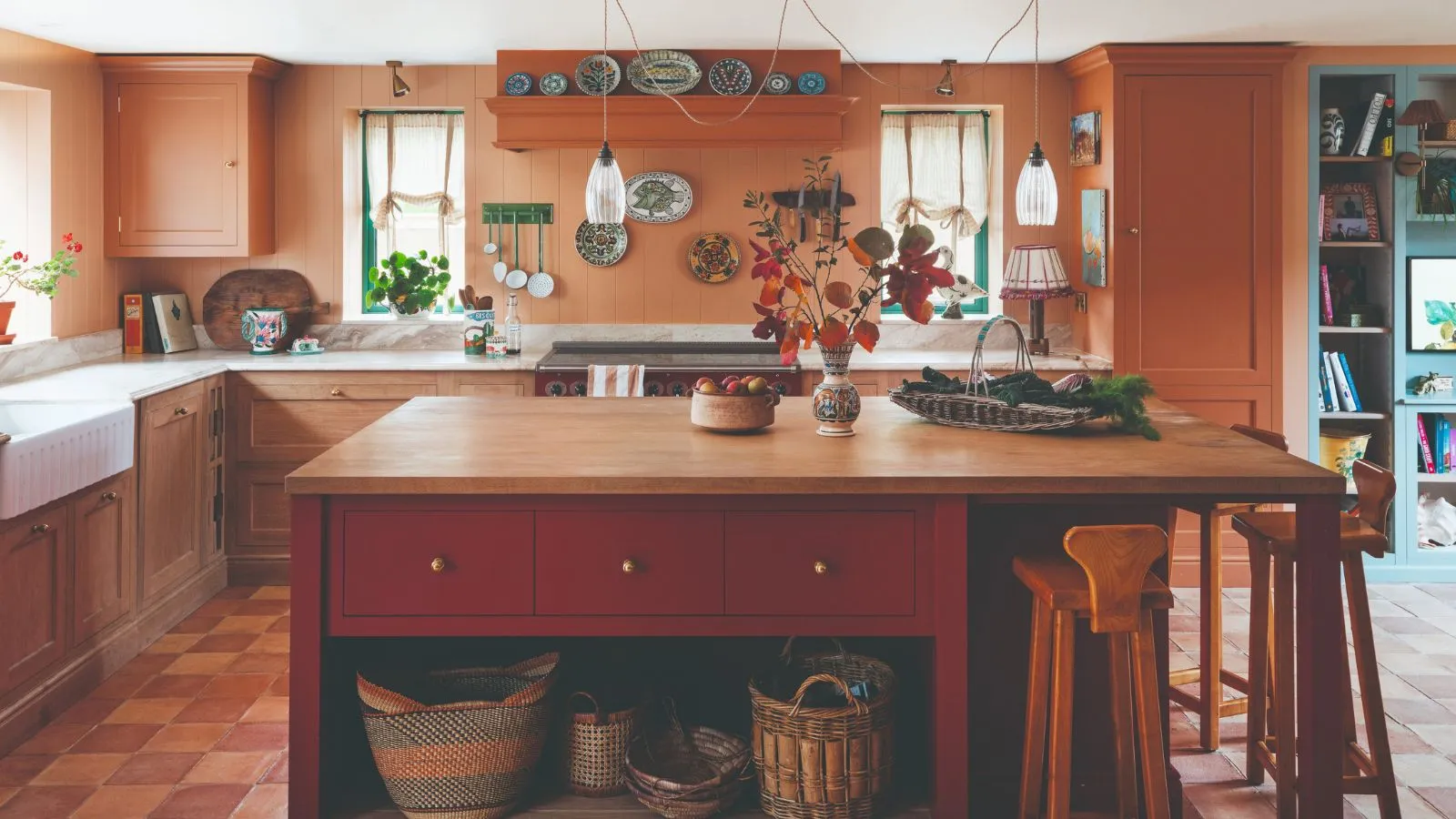 Tradycyjna kuchnia z pomalowaną na czerwono wyspą wolnostojącą z drewnianym blatem. Ściany pomalowane na jaśniejszy kolor czerwony z wystrojem wiejskim.' loading='lazy' title=