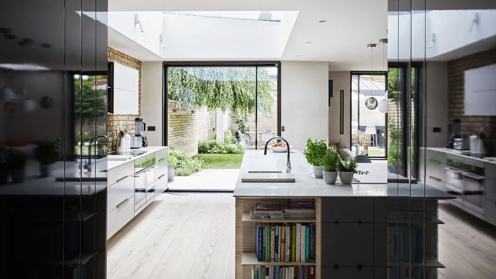 Jasna i przestronna, nowoczesna kuchnia z drewnianą podłogą, czarno-białymi szafkami, wyspą z blatami z białego marmuru i wbudowanymi półkami wypełnionymi książkami kucharskimi, odsłoniętym ceglanym tyłem i dużymi oknami prowadzącymi do bujnego, zielonego ogrodu.' loading='lazy' title=