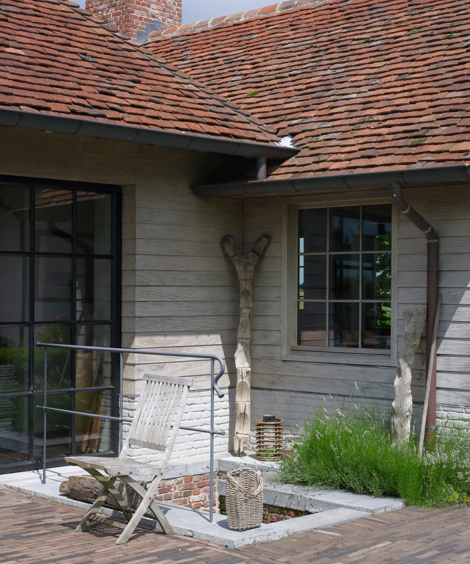 Dom wyłożony boazerią, z oknami Crittal, czerwonymi dachówkami, drewnianym krzesłem oraz rynną i rurą spustową.' loading='lazy' title=