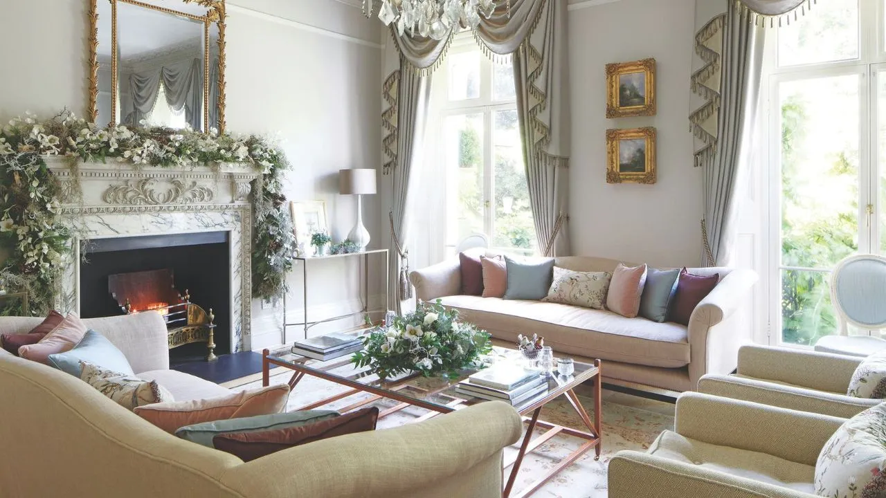 Elegancki salon z białymi ścianami, białymi i złotymi zasłonami z frędzlami nad dużymi oknami z tyłu pokoju i dużym marmurowym kominkiem po lewej stronie z rozpalonym ogniem, dużą girlandą z kwiatów i dużym złotym lustrem powyżej. Na środku pokoju znajduje się duży szklany stolik kawowy z kwiatami, a wokół niego wyściełane różowo-żółte kanapy i krzesła z poduszkami w różnych kolorach. Pod stolikiem kawowym znajduje się wzorzysty dywan.' loading='lazy' title=