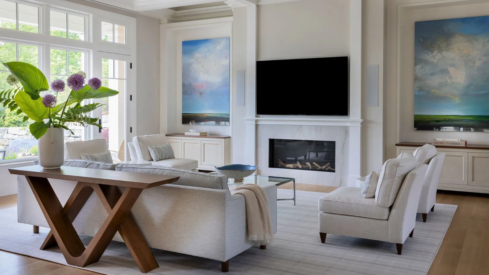 Weißes Wohnzimmer mit hoher Decke, Kamin, montiertem Fernseher über Hartböden und Teppich mit Sofas, die den Fernseher in der Mitte platzieren. Große Fenster sorgen für viel natürliches Licht und eine Holzkonsole im Vordergrund präsentiert riesige violette Blüten und große grüne Blätter' loading='lazy' title=