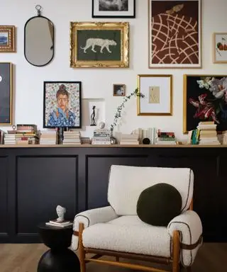 Mur de galerie rempli d'œuvres d'art dans des cadres et des miroirs de différentes tailles, accroché au-dessus d'un panneau latéral noir devant un fauteuil en boucle blanche avec un coussin noir