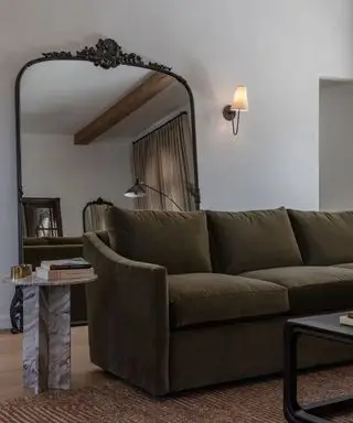 Grand miroir de sol antique posé contre le mur derrière un canapé vert olive avec une petite table d'appoint en marbre et une applique murale