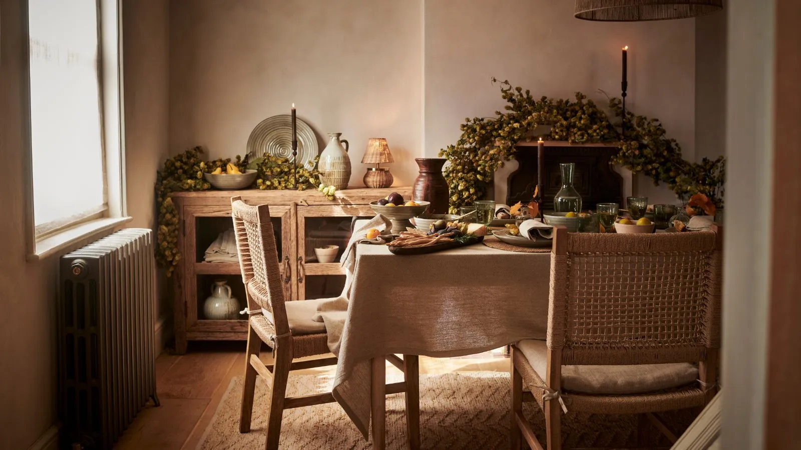 Rustykalna jadalnia z jesienną girlandą nad kominkiem. Prostokątny stół nakryty kremowym lnianym obrusem i podciągnięte do niego drewniane krzesła. W tle witryna z zastawą stołową w środku.' loading='lazy' title=