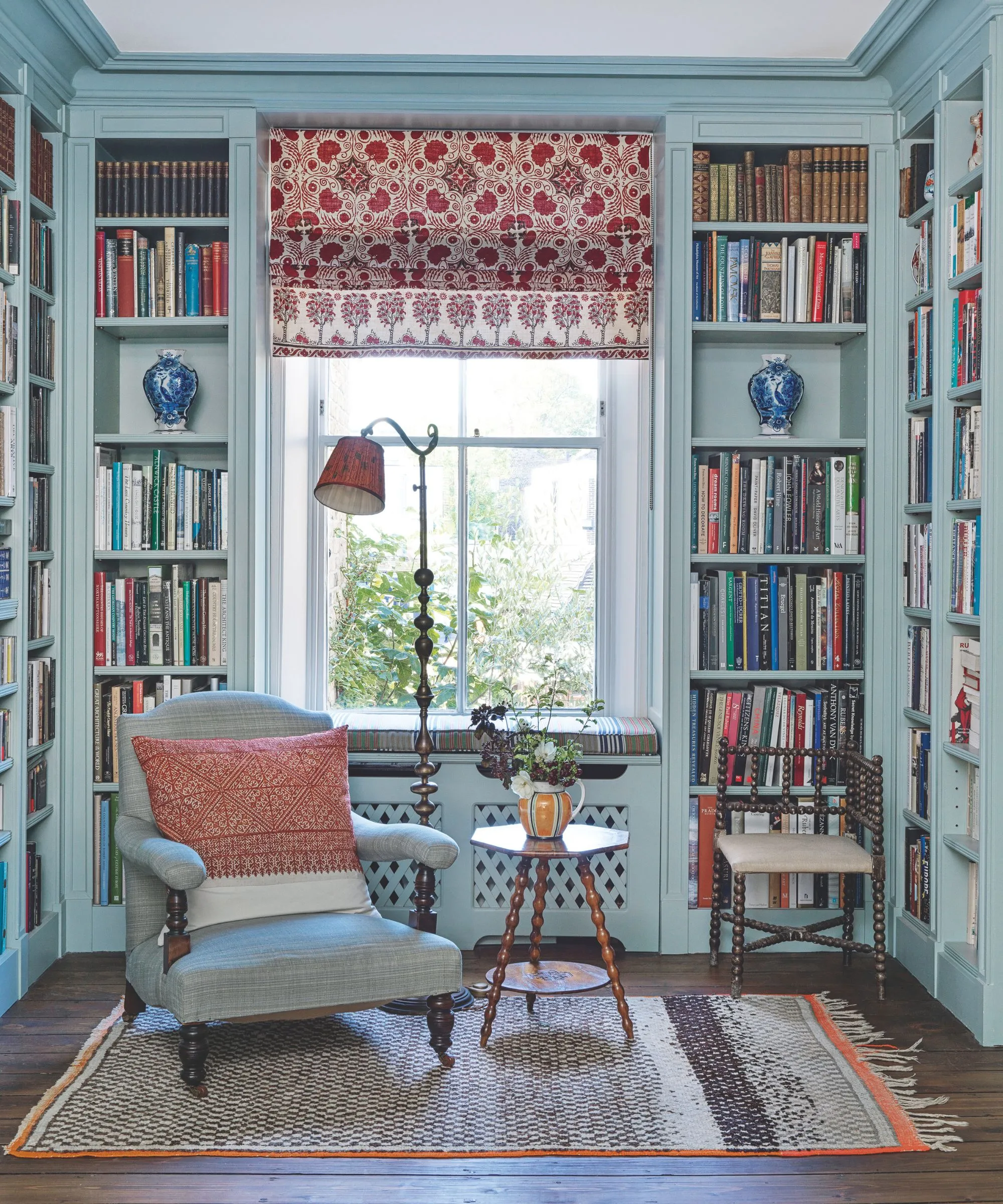 Niebieski salon z półkami na książki, niebieskim tapicerowanym krzesłem, drewnianą lampą podłogową i małym drewnianym okrągłym stołem. Za nim znajduje się duże okno z zasłoną w czerwono-biały wzór.' loading='lazy' title=