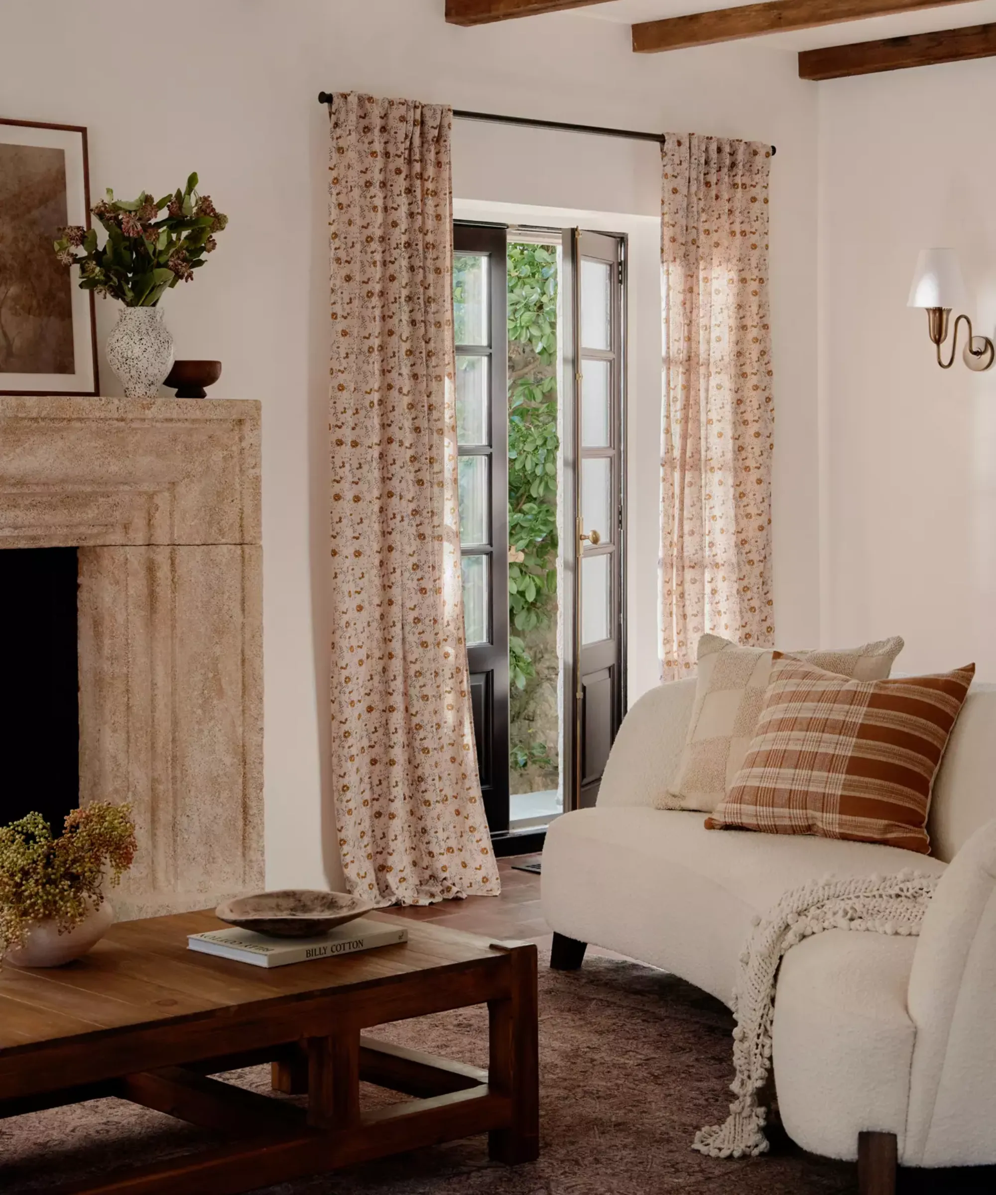 pokój dzienny z długimi kwiatowymi zasłonami nad francuskimi drzwiami, kanapą boucle z poduszkami, drewnianym stolikiem kawowym i kominkiem' loading='lazy' title=
