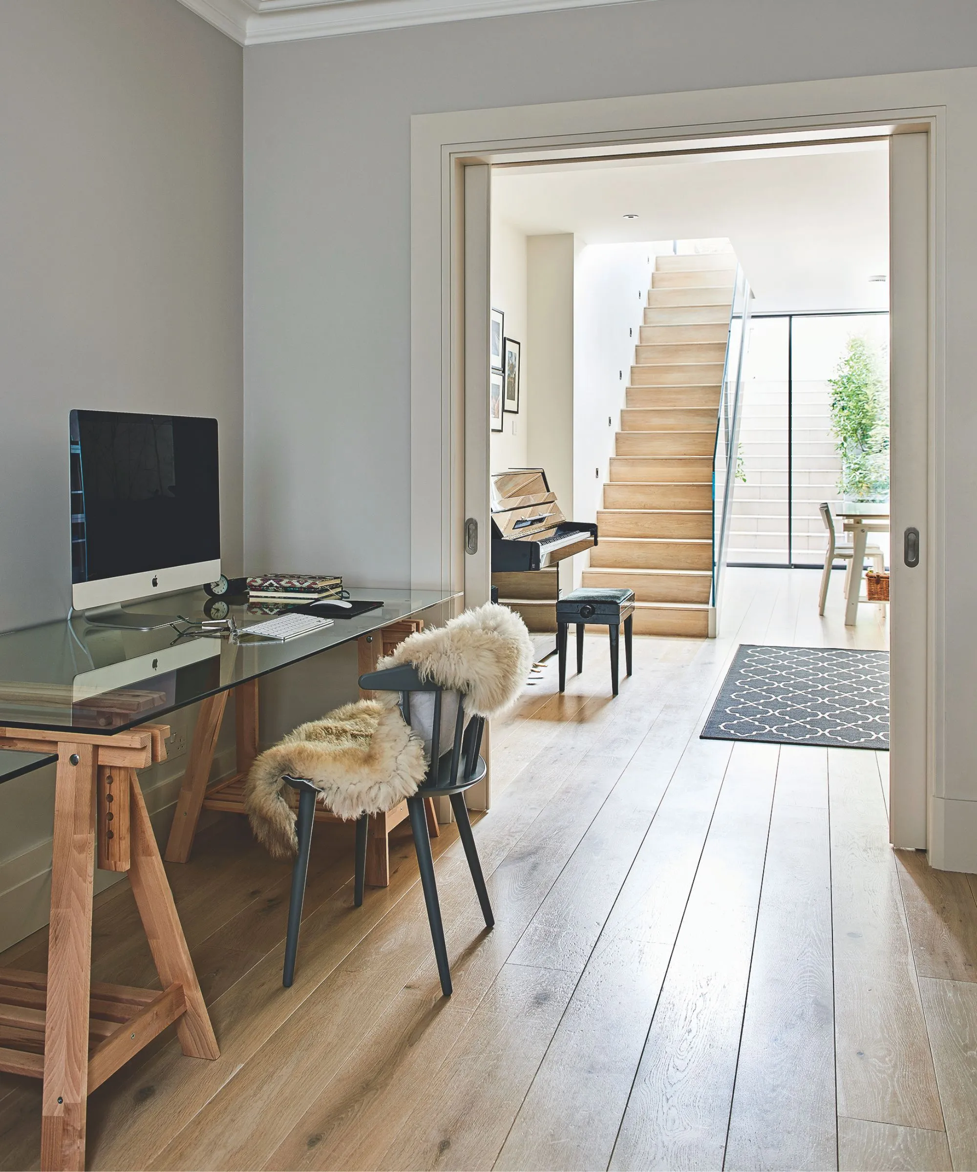 En lys og luftig kælder med trægulv, et skrivebord i træ og glas med en træstol med et blødt kast og et klaver i baggrunden tilstødende værelse med et sort mønstret tæppe og en trætrappe ovenpå. I baggrunden ses glasdøre ud til en havetrappe med en bladgrøn plante til højre.' loading='lazy' title=