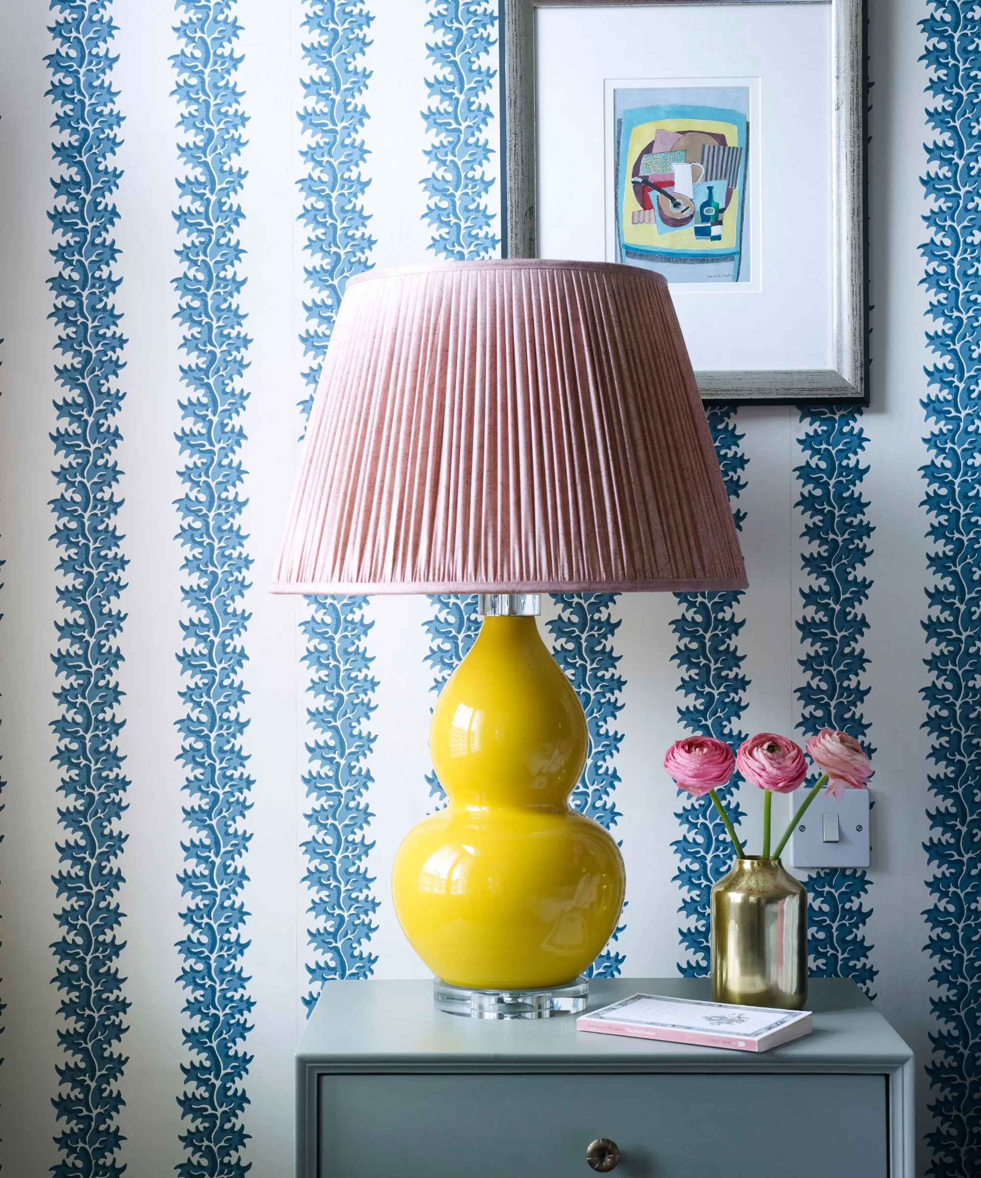 Et værelse med blåt og hvidt statement tapet en gul keramisk lampe med lyserød lampeskærm indrammet kunstværk i sølvramme og blåt sengebord med en guldvase fyldt med lyserøde blomster og en bog.' loading='lazy' title=