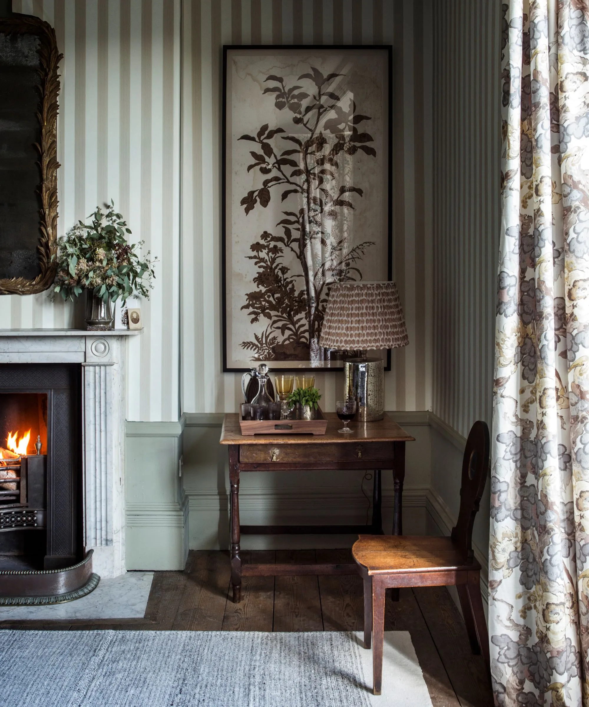 En hyggelig høstlig stue med stribet tapet et oplyst brandkunstværk blomstergardiner og et stort guldindrammet spejl. Gulvet er træ med et gråt tæppe på toppen.' loading='lazy' title=