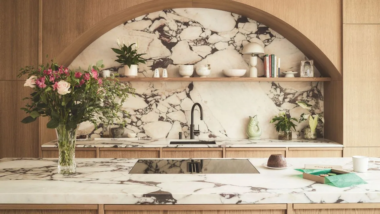 Nowoczesna kuchnia z łukowymi okuciami z jasnego drewna nad zlewem kuchennym z dużym marmurowym tyłem. Wyspa naprzeciwko z dopasowanym marmurowym blatem i szklaną płytą kuchenną. Po lewej stronie wazon z różami.' loading='lazy' title=