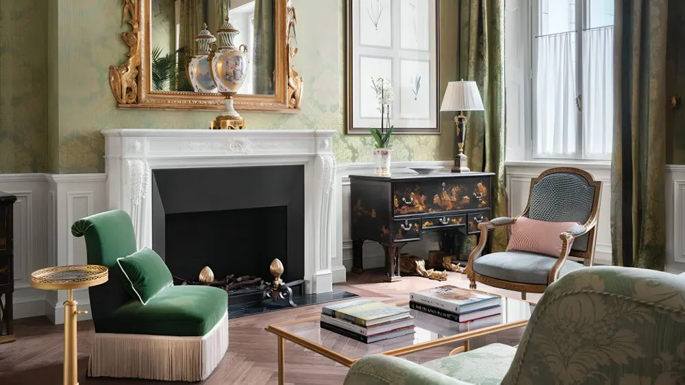 Tradycyjny salon z zieloną tapetą, dużym kominkiem, szklanym stolikiem kawowym i zielonymi fotelami.' loading='lazy' title=