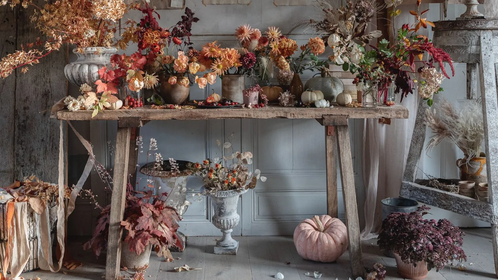 Una stanza con pannelli grigi e un tavolo in legno vintage pieno di fiori di Halloween e decorazioni tra cui zucche' loading='lazy' title=