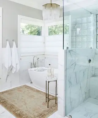Espace salle de bain blanc et gris avec carrelage en marbre au sol et sur les murs, tapis neutre et texturé, douche avec porte en verre, suspension en verre et or, serviettes sur crochets au mur, fenêtres avec stores blancs, table d'appoint élancée en métal positionnée à côté au bain blanc