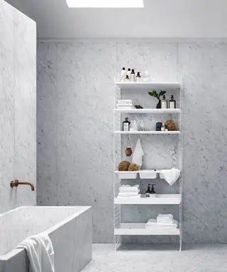 Salle de bains minimaliste et moderne avec étagères à cordes blanches, décorée avec des serviettes, des articles de toilette, des objets décoratifs, des carreaux de marbre assortis sur les murs et les sols, une baignoire carrelée assortie avec un robinet en cuivre