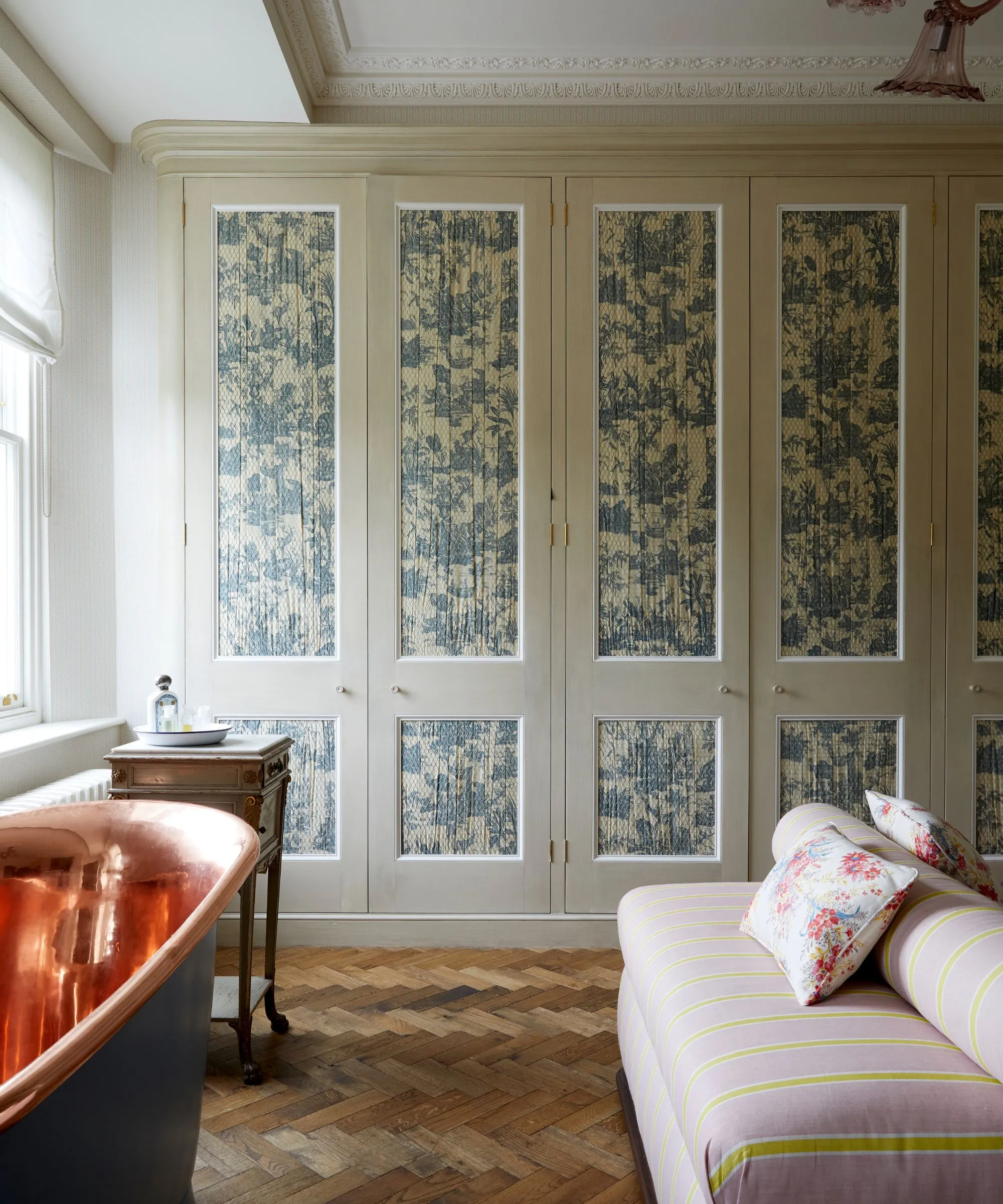 Um armário estampado com piso em parquet de madeira, uma mesa de banheira de madeira cobre e preta e um sofá listrado rosa e amarelo com almofadas florais.' loading='lazy' title=