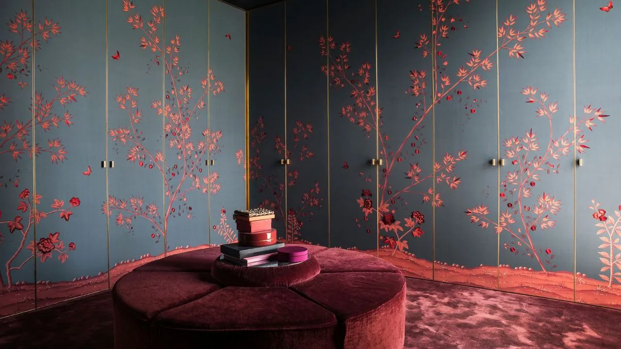 Closet de luxo escuro com guarda-roupas embutidos do chão ao teto, portas com papel de parede azul escuro fosco e design floral oriental vermelho. Tapetes de pelúcia cor de vinho com veludo redondo combinando com assento grande no centro' loading='lazy' title=