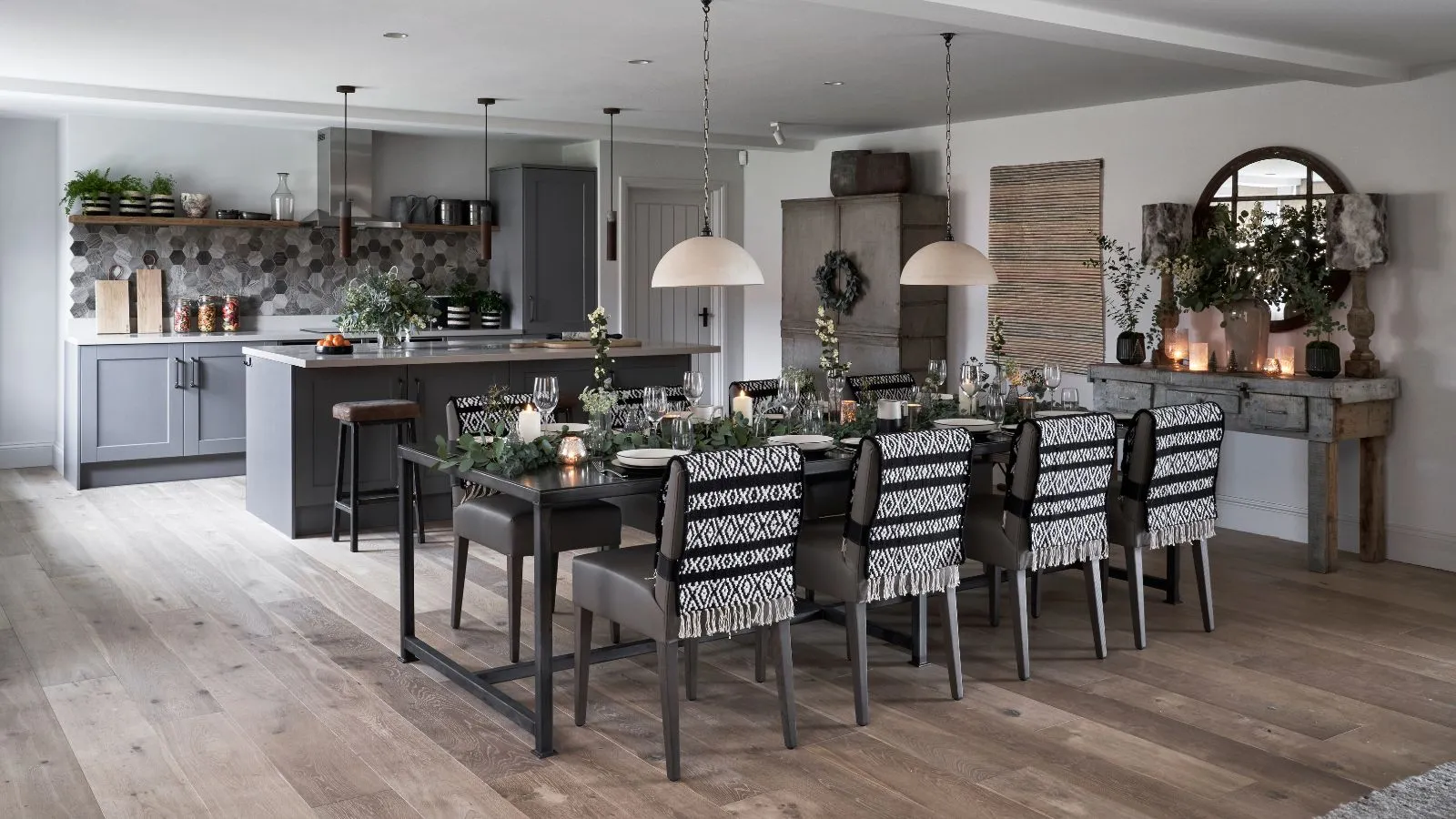 Czarno-biało-szara kuchnia z szarymi szafkami i drewnianymi podłogami. W tle szara wyspa kuchenna, a z przodu duży, nakryty stół jadalny pokryty gałęziami, zapalone świece w szklankach, a na krzesłach rozłożone czarno-białe koce. Po prawej stronie znajduje się okrągłe lustro. Ściany są białe.' loading='lazy' title=