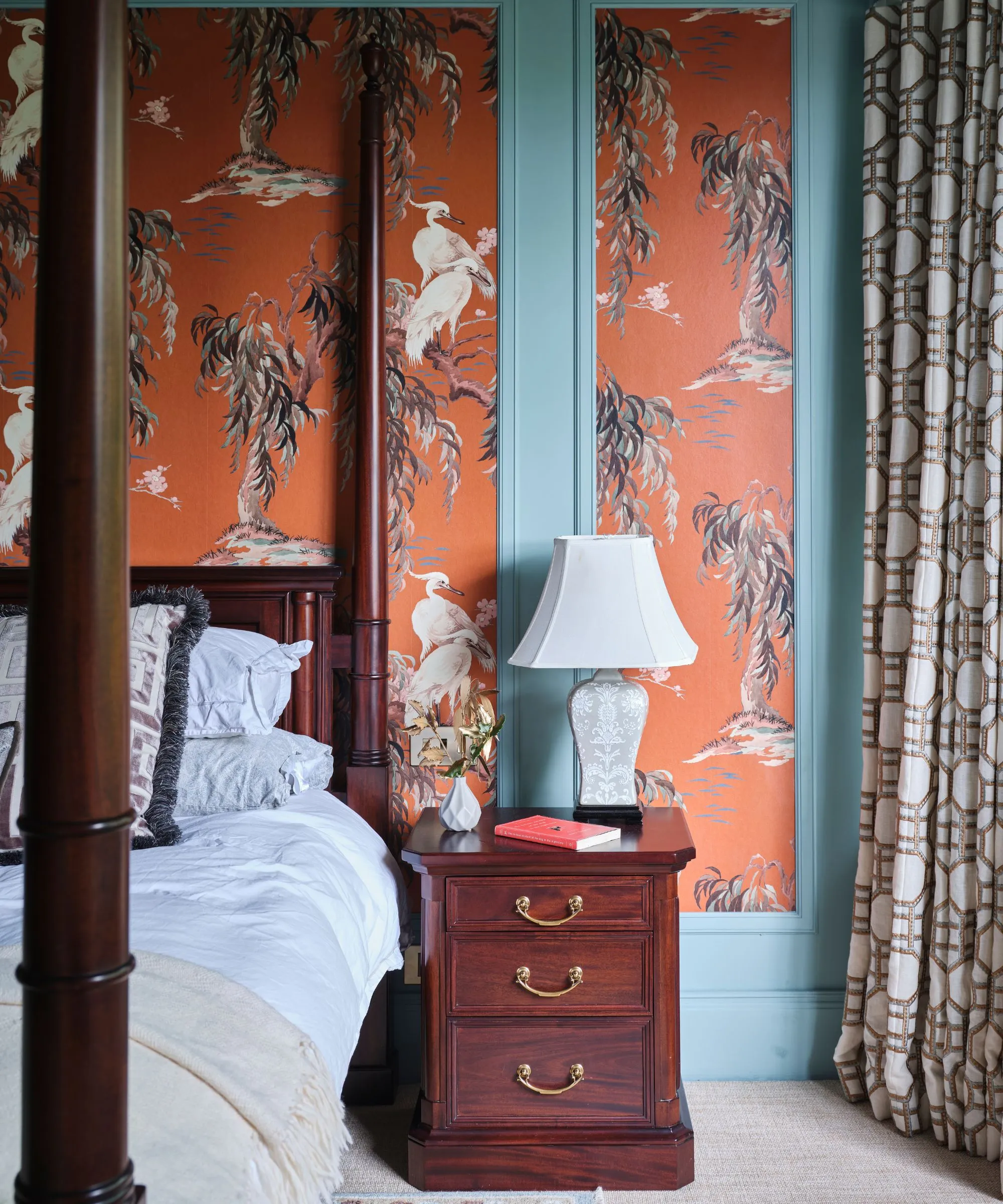 Sypialnia z niebieskimi ścianami i pomarańczową tapetą z panelami, łóżkiem z baldachimem z ciemnego drewna, białą pościelą, pasującym stolikiem nocnym, zasłonami we wzór białych lamp i neutralnym dywanem.' loading='lazy' title=
