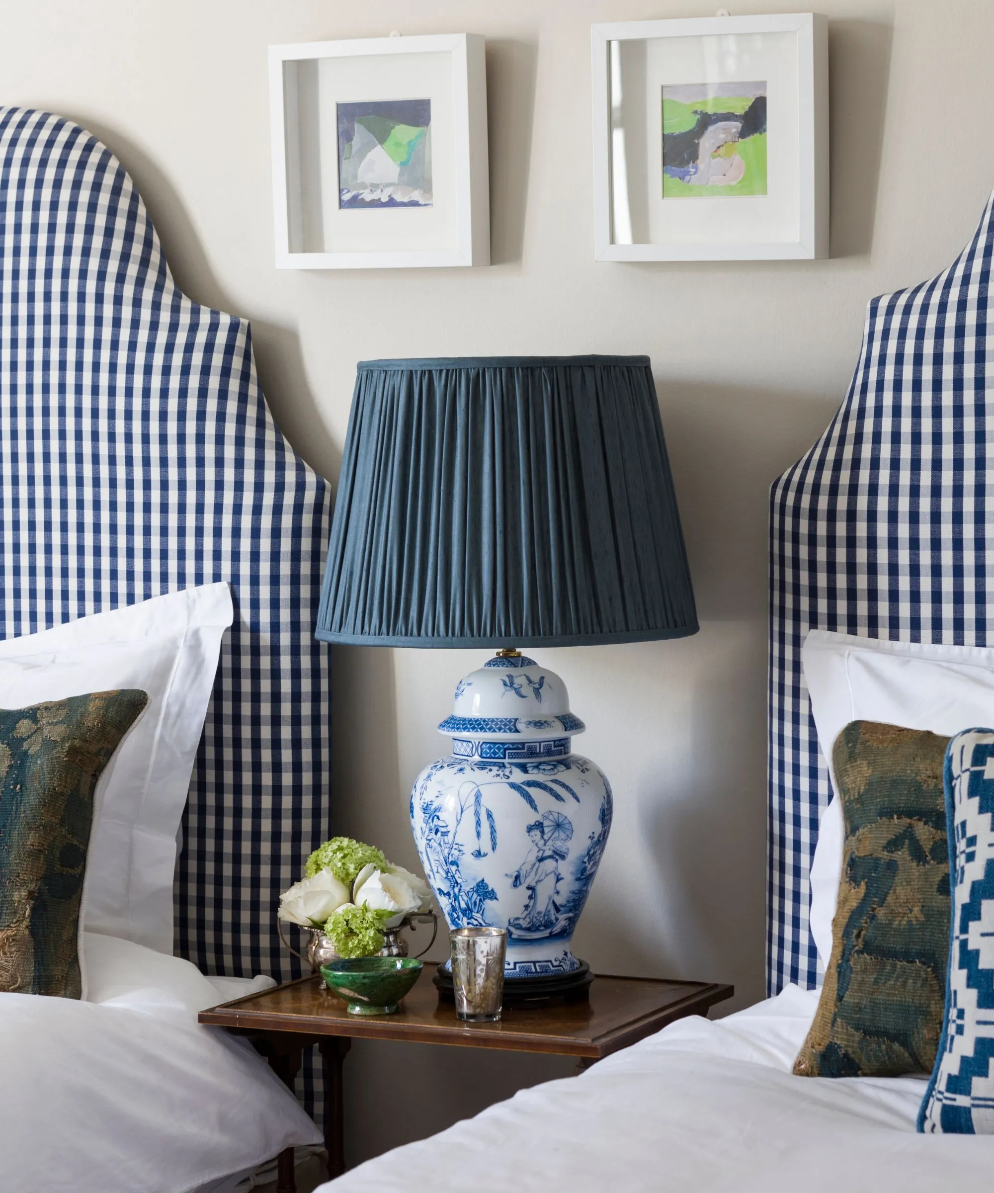 Biała sypialnia z dopasowanymi podwójnymi łóżkami z zagłówkami w kratkę i brązowym drewnianym stolikiem nocnym pomiędzy nimi. Na stoliku nocnym stoi ceramiczna lampa dekoracyjna z turkusowym abażurem, a nad nią dwie prace oprawione w ramy.' loading='lazy' title=