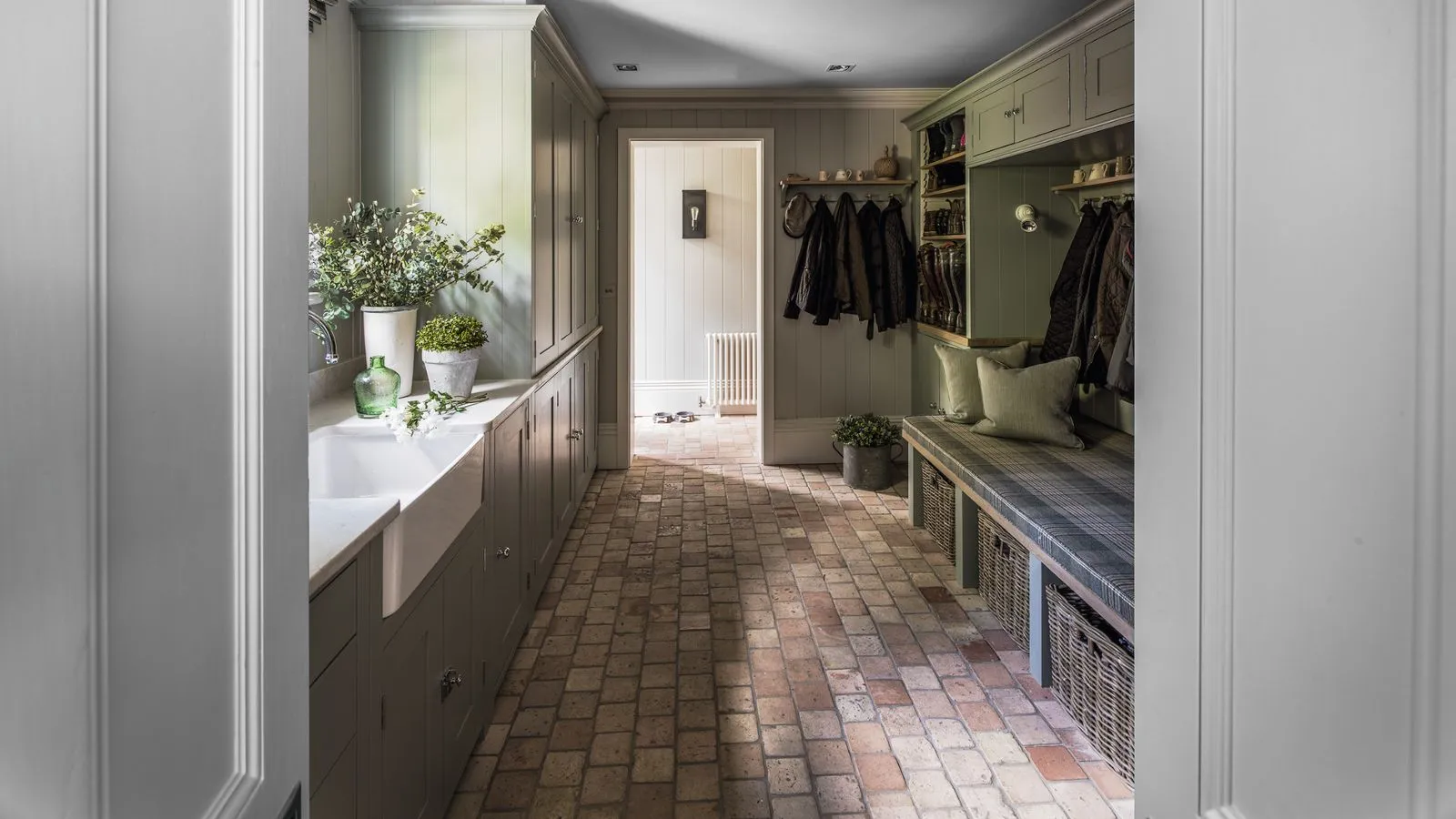 Uno stivale piastrellato con armadietti verde salvia, cesti di vimini, una panca di legno con cuscini verdi, un lavandino bianco e piante d'appartamento verdi in vasi bianchi.' loading='lazy' title=