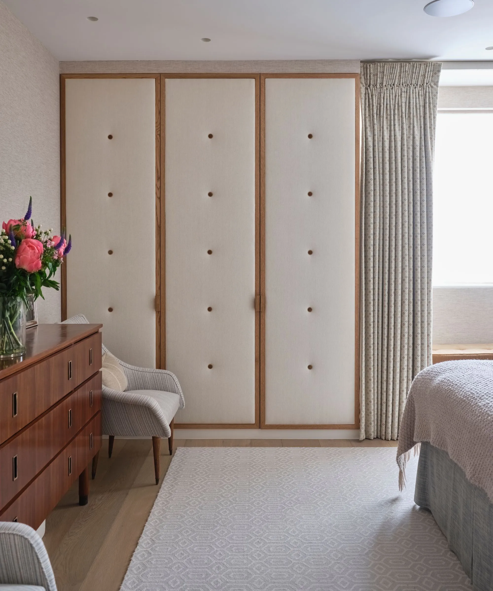 Un armadio imbottito color crema e legno in una camera da letto con un tappeto color crema, una credenza in legno con un vaso di fiori rosa brillante e una sedia grigia.' loading='lazy' title=