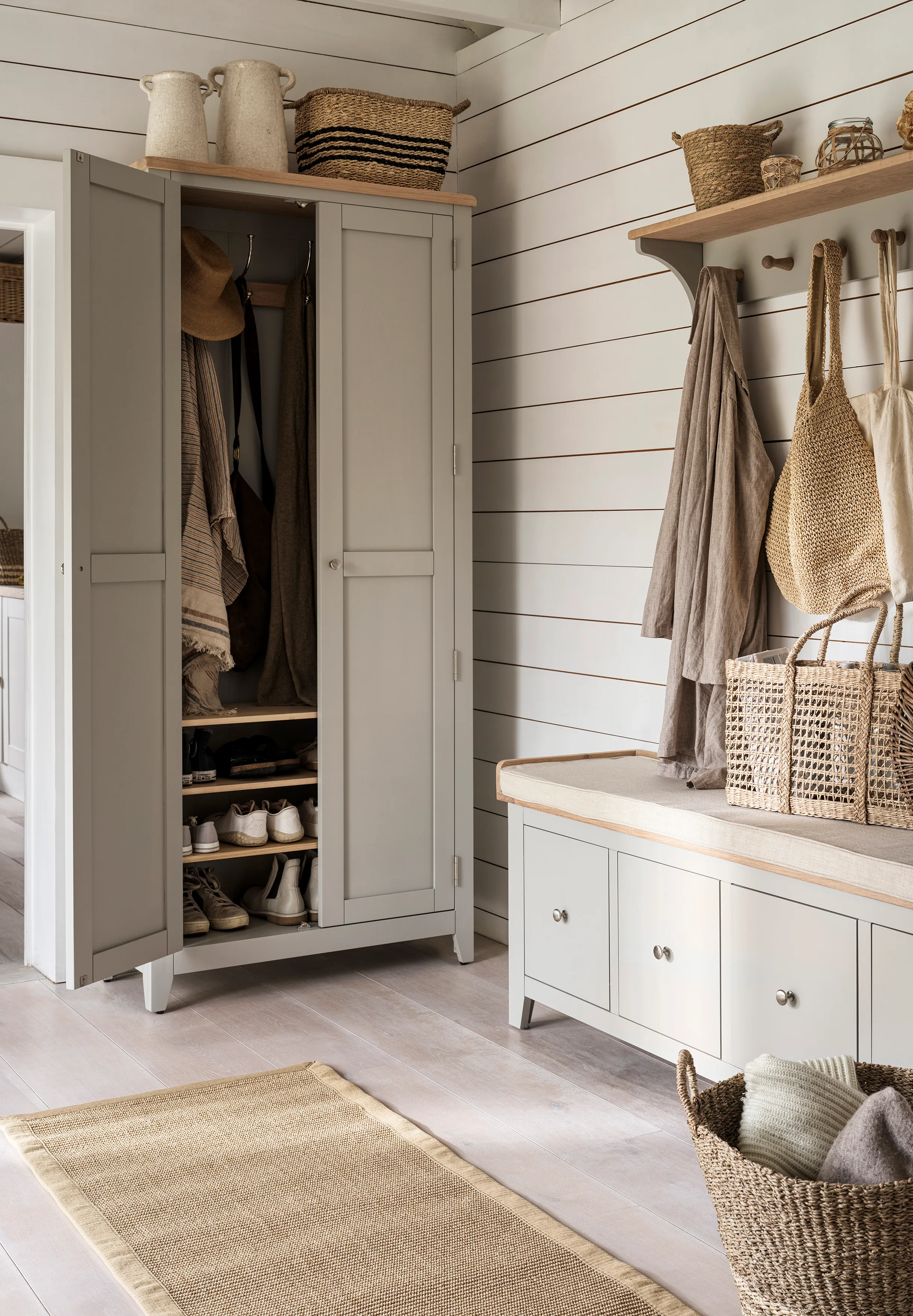 Un armadio autoportante verniciato di grigio in un ingresso bianco sul bordo di una nave accanto a una panca per scarpe abbinata con cassettiera e un appendiabiti a muro con cappotti appesi' loading='lazy' title=
