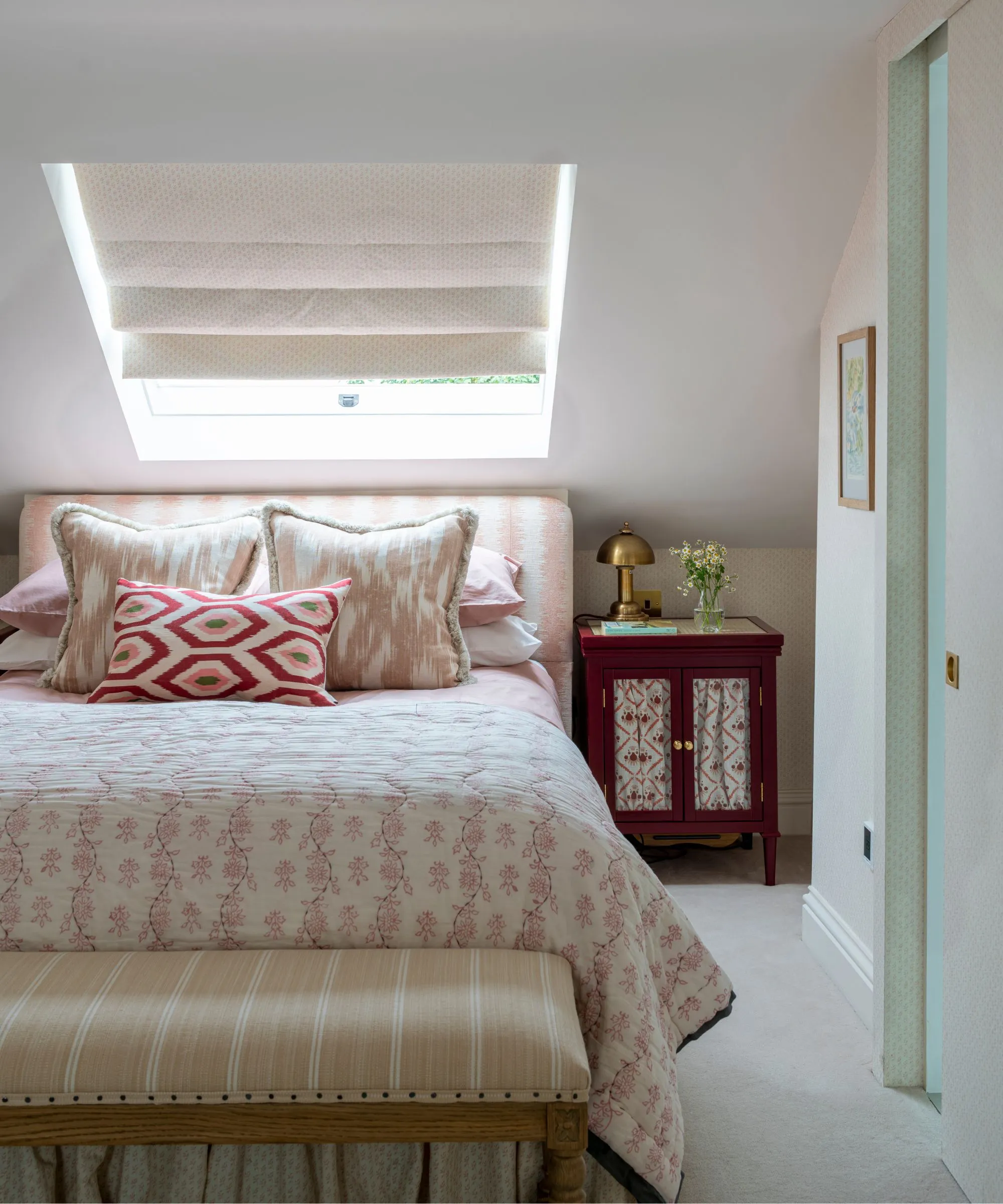 Una camera da letto mansardata bianca con un grande letto matrimoniale con cuscini e testiera a motivi rosa e bianchi, un comodino marrone ottomano a righe con lampada dorata e tenda bianca su una finestra inclinata sopra il letto.' loading='lazy' title=