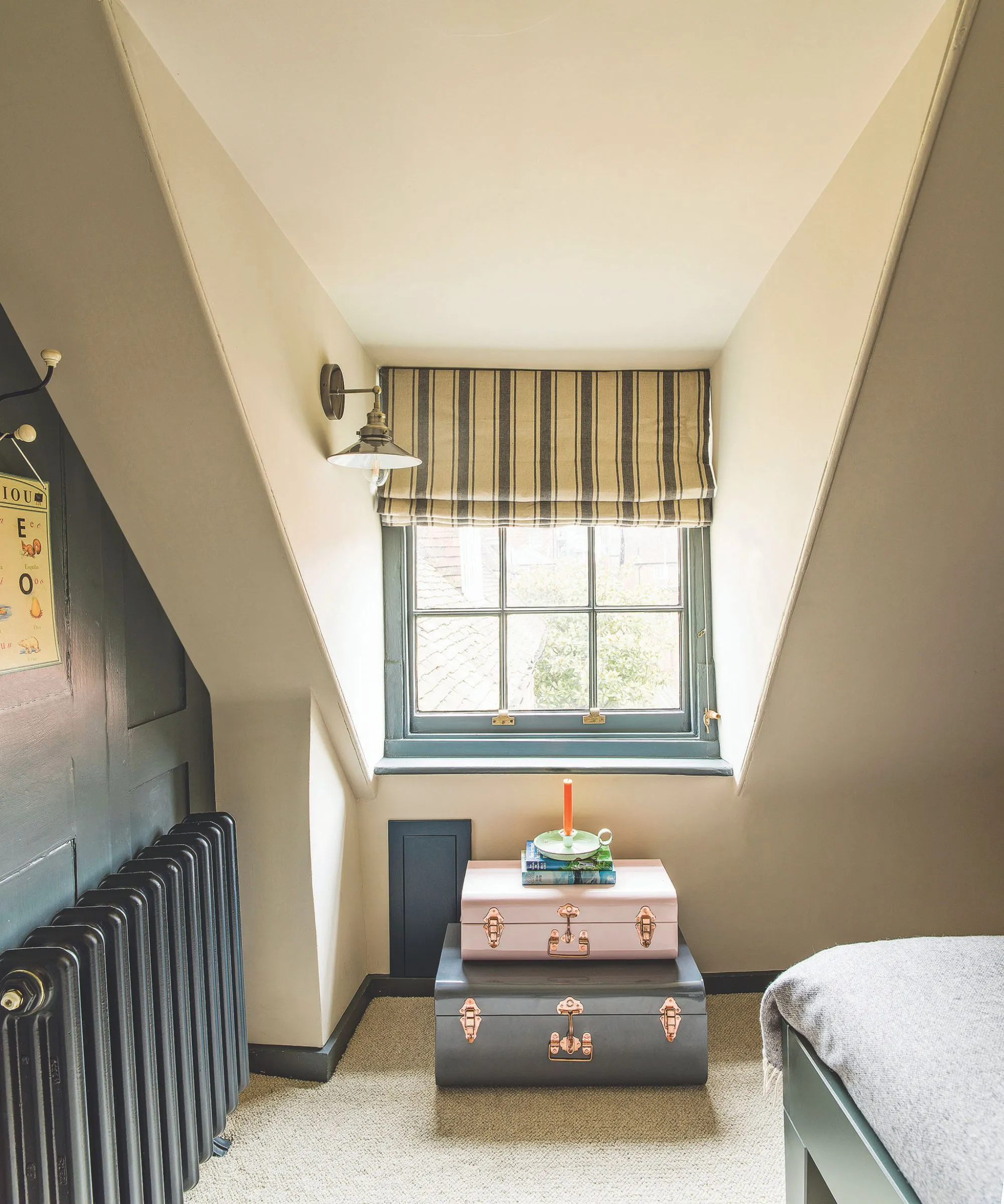 Białe poddasze z turkusową ścianą i grzejnikiem, roletą w białe i niebieskie paski oraz zestawem walizek. Po prawej stronie znajduje się róg niebieskiej ramy łóżka z szarą pościelą.' loading='lazy' title=