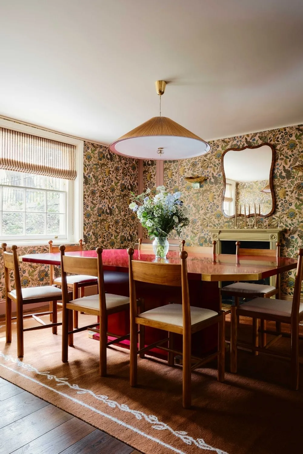 květinově tapetovaná jídelna s velkým červeným leskem lakovaným stolem dřevěné starožitné židle s míčem s detaily velkého koberce s detaily okraje a krbem a zrcadlem' loading='lazy' title=