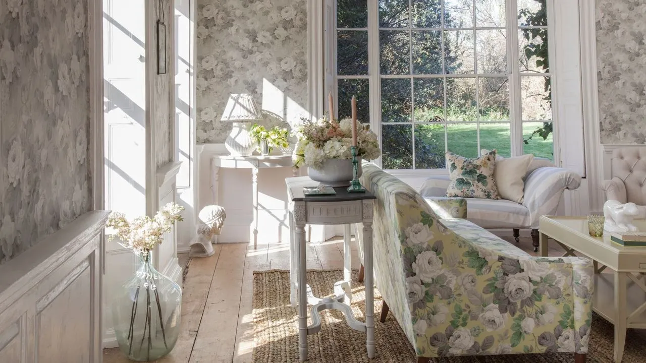 Kwiatowy salon z kwiecistą tapetą Kwiecista kanapa, biały stolik kawowy, rattanowy dywan, drewniana podłoga, duży szklany wazon z białymi kwiatami i dużymi oknami wychodzącymi na pełen zieleni ogród.' loading='lazy' title=