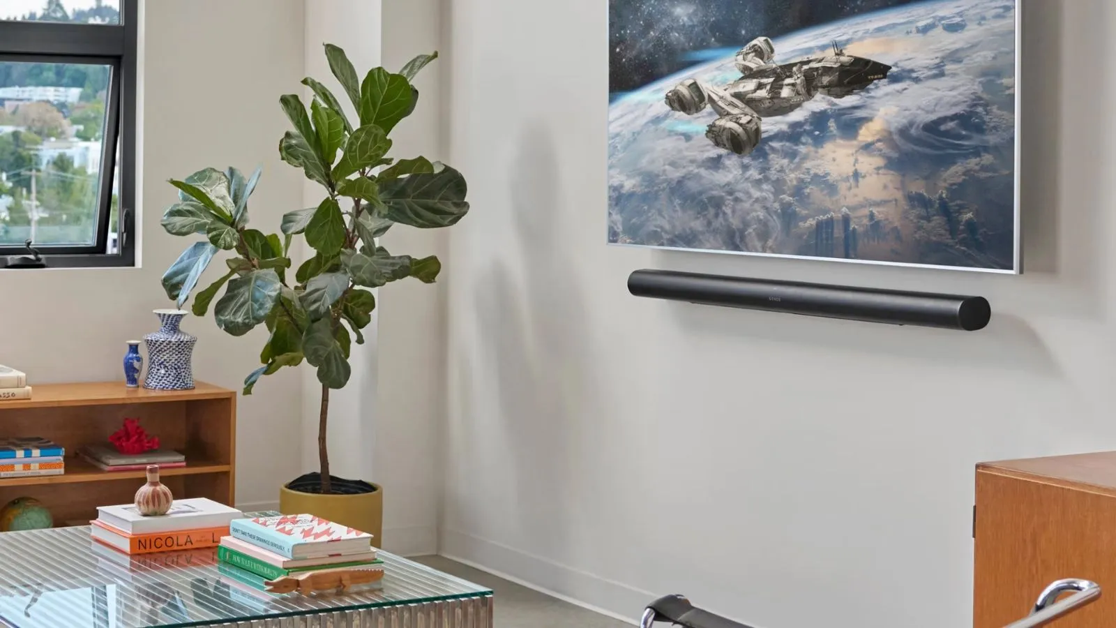 Sonos Arc Ultra sotto la TV che mostra una scena spaziale in un soggiorno bianco con tavolino in vetro vegetale, libreria in legno e finestra parzialmente aperta.' loading='lazy' title=