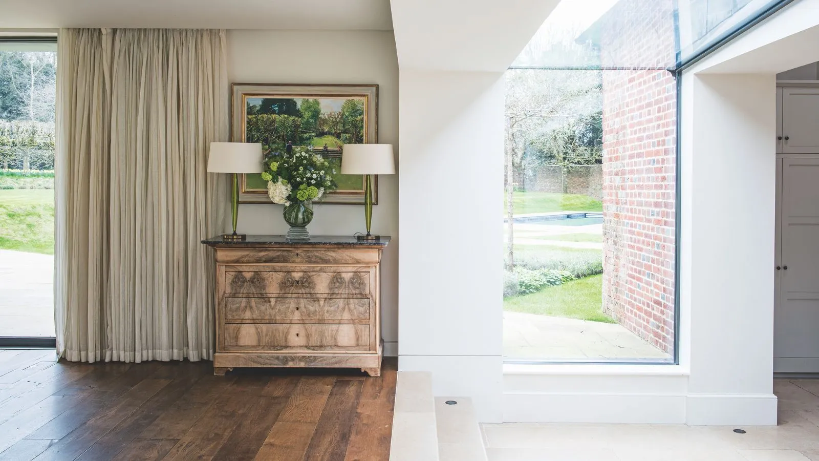 Jasna przestrzeń przejściowa ze szklanymi oknami, drewnianą podłogą, białymi ścianami i brązową komodą z dwiema pasującymi lampami z białymi kloszami, grafiką powyżej i kremowymi zasłonami po lewej stronie.' loading='lazy' title=