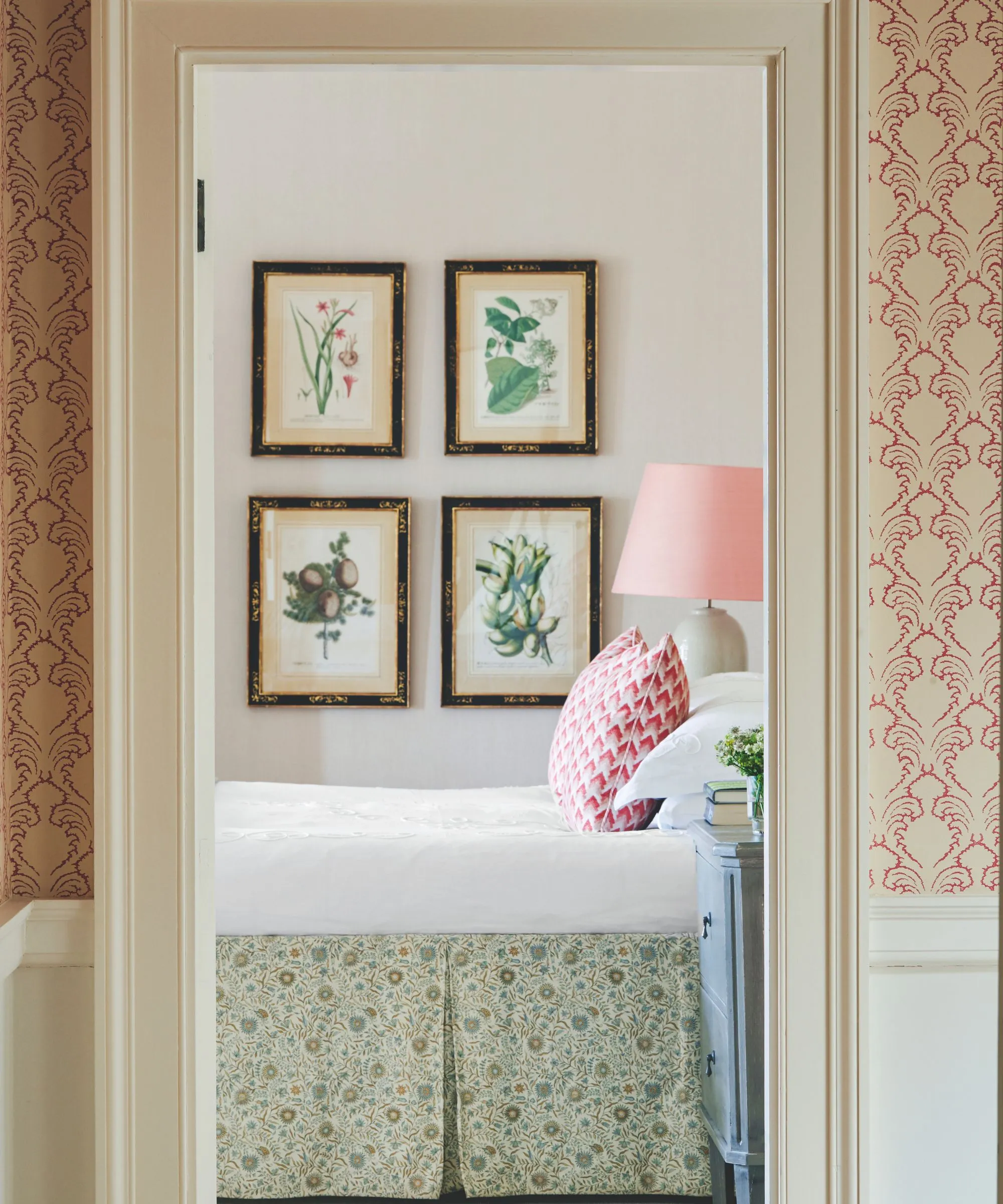 Białe drzwi z różową i białą tapetą po obu stronach i białymi listwami przypodłogowymi ukazującymi widok na sypialnię z łóżkiem z białą pościelą, zieloną kwiecistą spódnicą i różowo-białą poduszką. Ściany są jasnoróżowe, a na ścianie znajduje się galeria z motywem kwiatowym. Obok łóżka na pierwszym planie znajduje się niebieski stolik nocny, a w tle biała ceramiczna lampa z różowym abażurem.' loading='lazy' title=