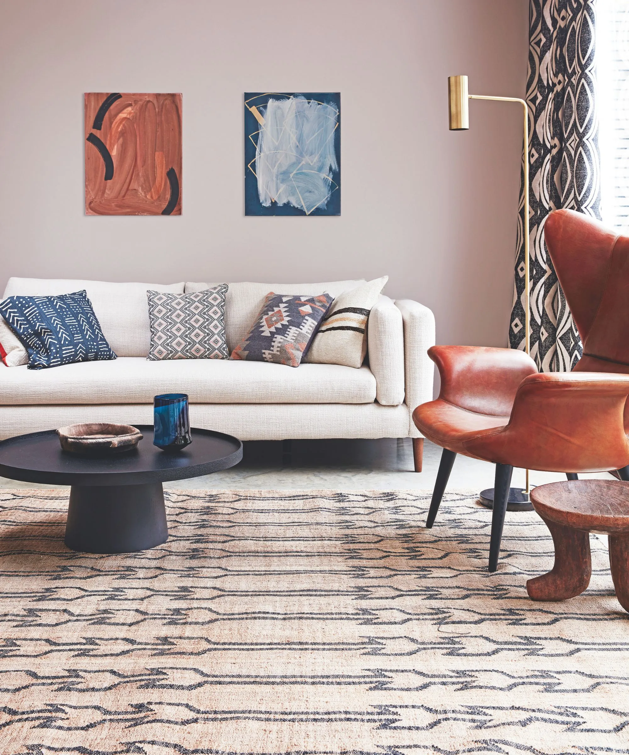 Pokój dzienny z wzorzystym dywanikiem, czarnym okrągłym stolikiem kawowym z miską i niebieskim wazonem na blacie, białą kanapą z wzorzystymi poduszkami, brązowym skórzanym krzesłem i drewnianym stołem obok niego. Z tyłu po prawej stronie znajduje się okno z wzorzystymi oknami i złotą lampą podłogową. Nad sofą znajdują się dwie abstrakcyjne dzieła sztuki.' loading='lazy' title=