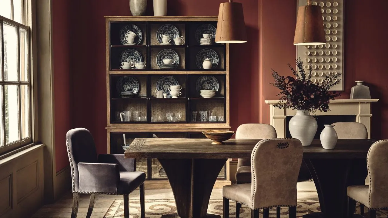 Ein tiefrotes Esszimmer mit einem großen Esstisch aus Holz. Drumherum gepolsterte Stühle und darüber zwei hängende braune Lederanhänger. Im Hintergrund ein Holzschrank mit feinem Geschirr.' loading='lazy' title=