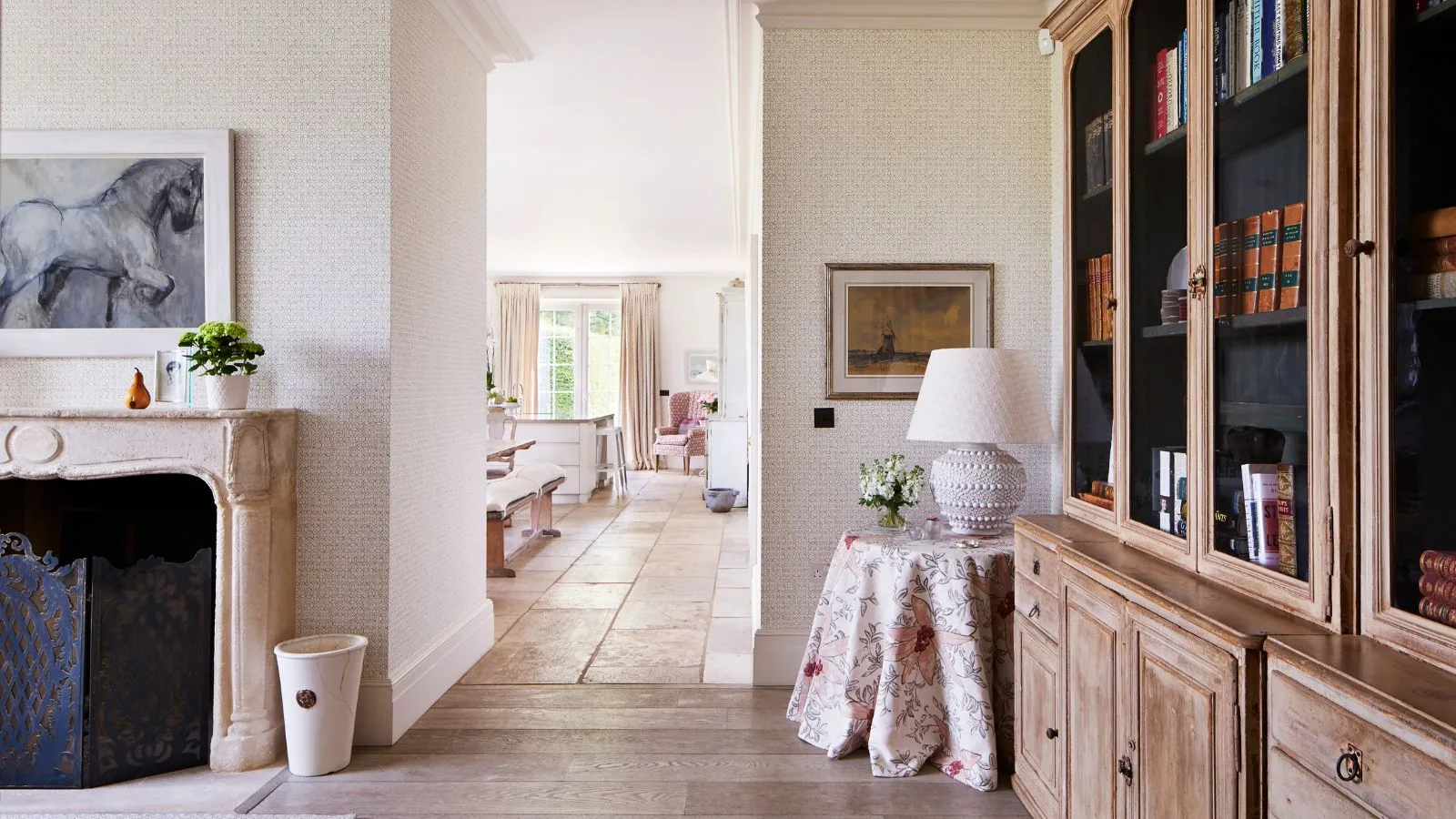 Jasny salon z białymi ścianami, drewnianymi szafkami i kamiennym kominkiem z grafiką konia powyżej. Za otwartym korytarzem widać kuchnię z ławami do siedzenia i wyspą kuchenną.' loading='lazy' title=