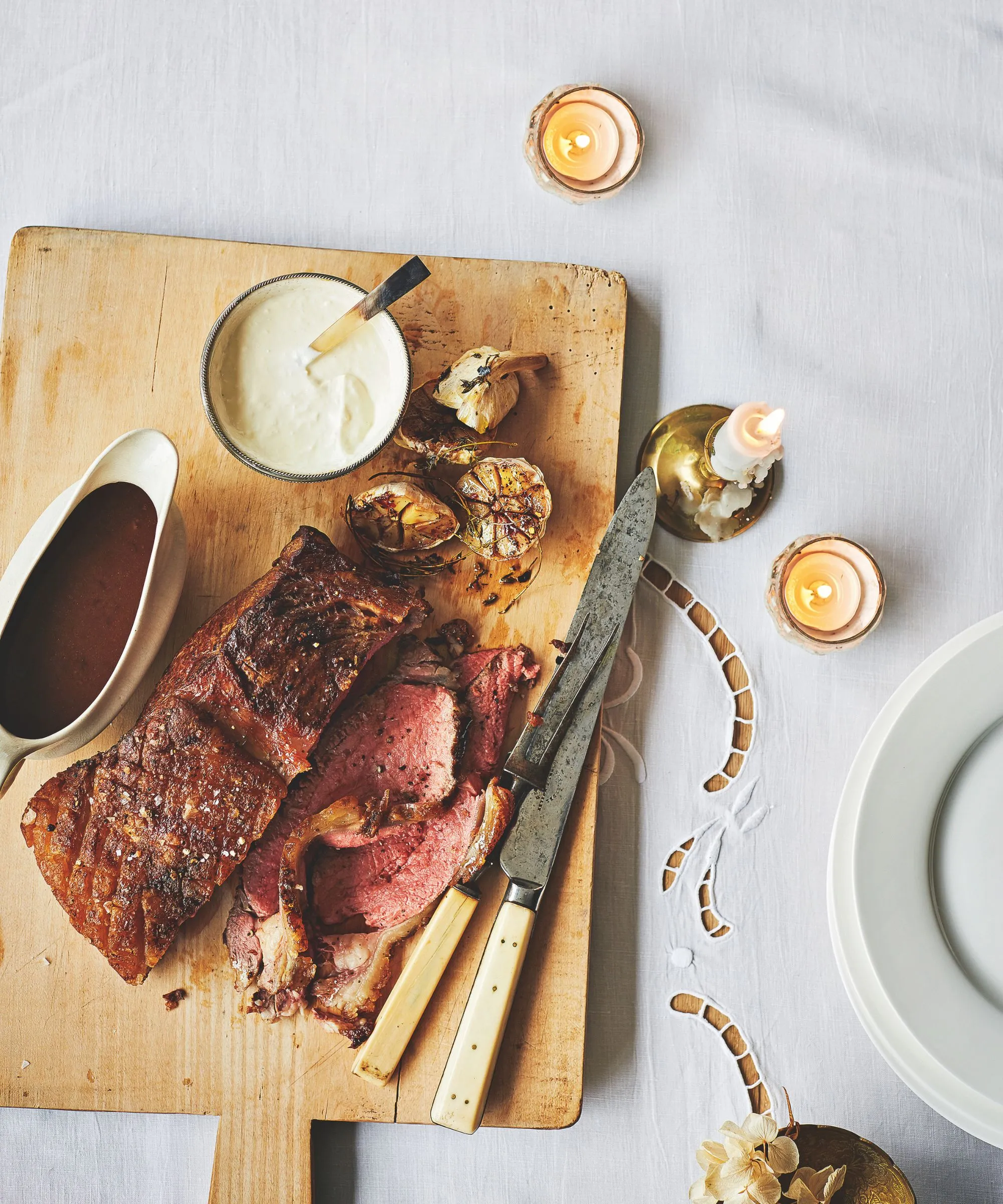 Pieczone mięso na drewnianej desce do krojenia na białym obrusie z białym nożem i widelcem do rzeźbienia sosu, świecami oraz stosem białych talerzy.' loading='lazy' title=