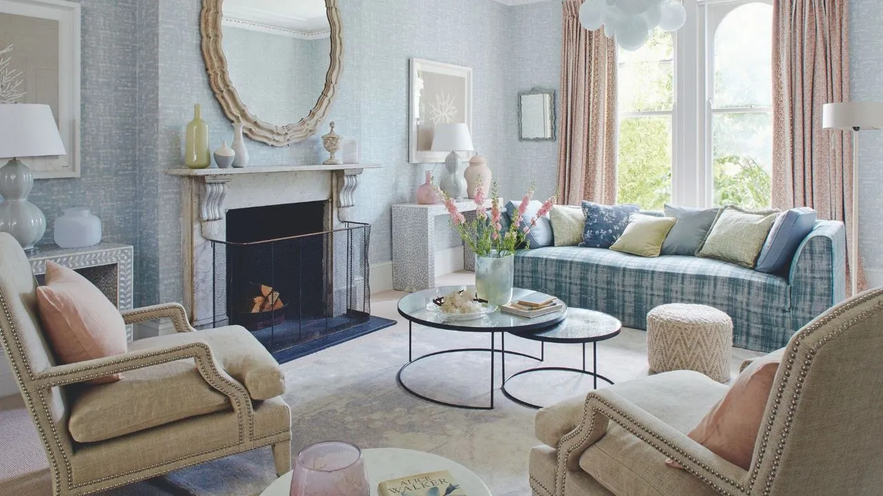 Pastelowy salon z tapetą w niebieskie wzory, kominkiem z białego marmuru z okrągłym lustrem powyżej i dużymi oknami po prawej stronie z pastelowymi różowymi zasłonami. Na pierwszym planie znajdują się dwa kremowe tapicerowane krzesła, a pośrodku pokoju znajduje się okrągły stolik kawowy z różowymi kwiatami, za którym stoi niebieska sofa w kratę. Na podłodze leży biały aksamitny dywan.' loading='lazy' title=