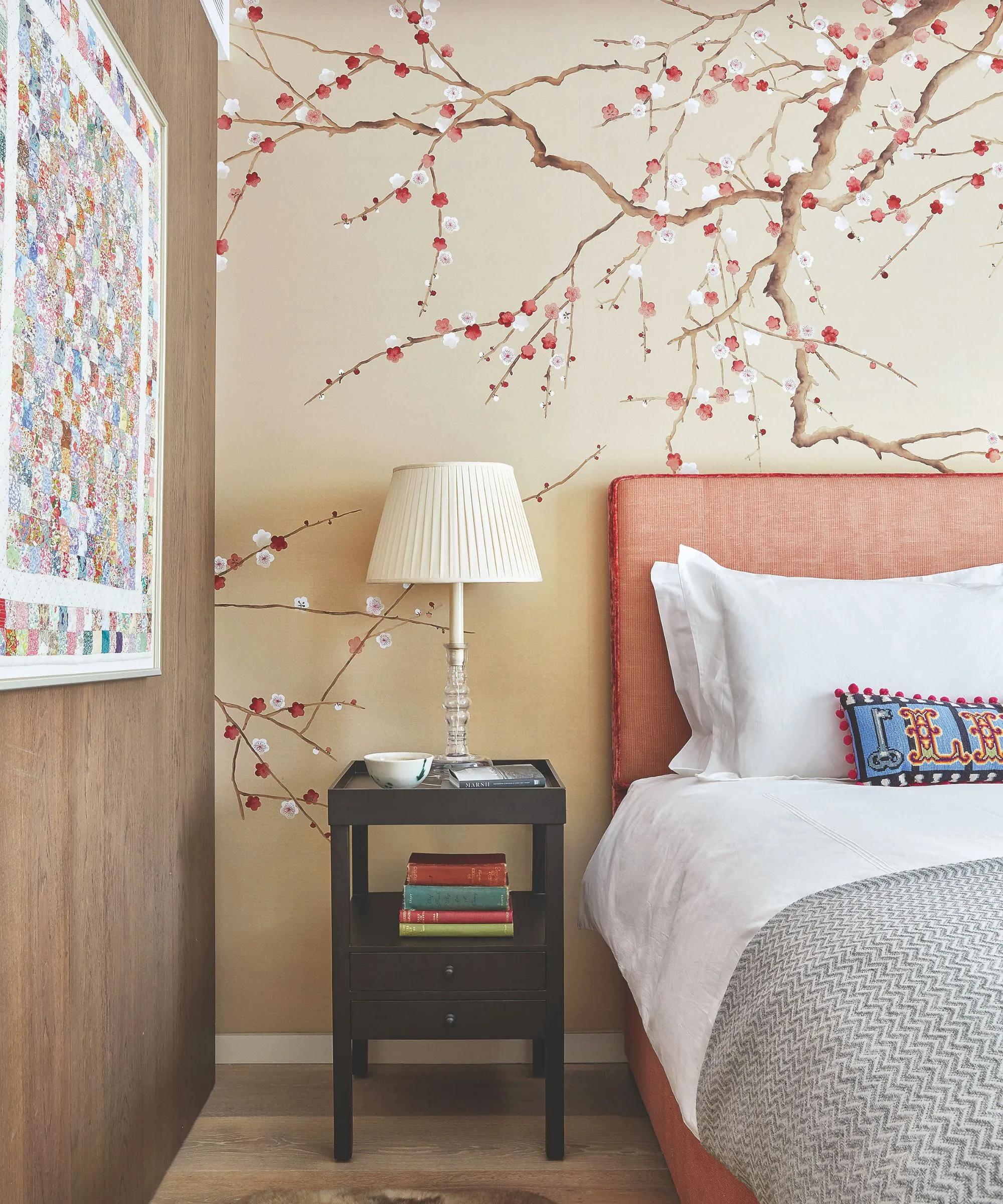 Sypialnia z pomarańczowym łóżkiem po prawej stronie z białą pościelą, szarym kocem i kolorową poduszką. Ściana za łóżkiem jest w neutralnym beżowym kolorze z motywem drzewa wiśni. Na lewo od łóżka znajduje się czarny stolik nocny ze stosem książek i białą lampą, a na lewo od niego ściana wyłożona boazerią z jasną i kolorową abstrakcyjną grafiką. Podłoga jest drewniana.' loading='lazy' title=