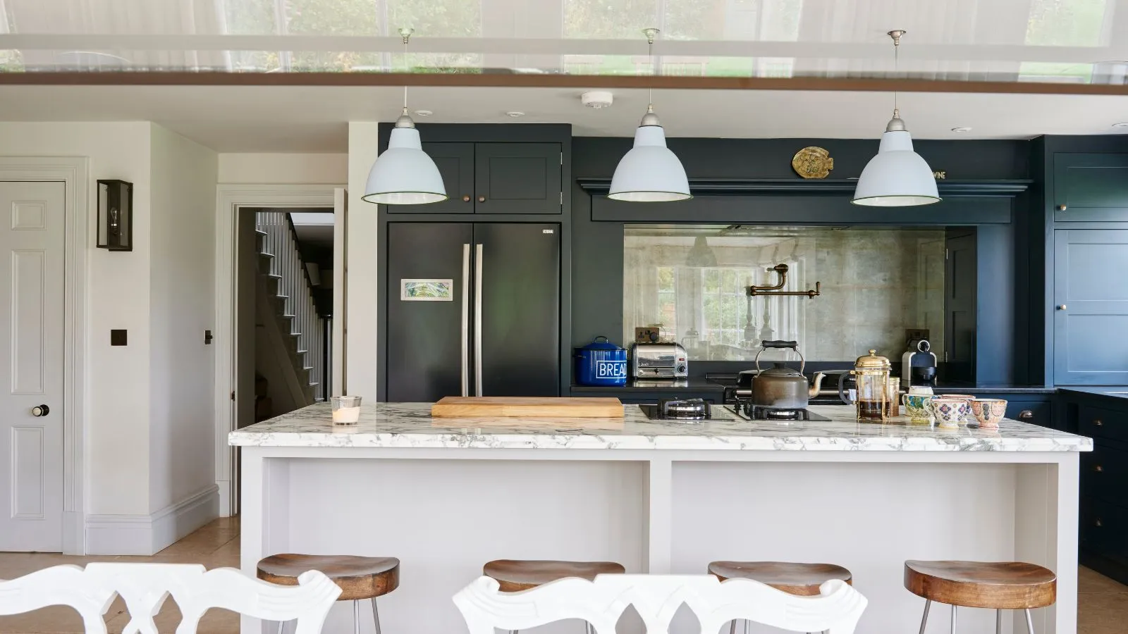 Eine helle und luftige Küche mit einer weißen Marmorinsel vier hölzerne Barhocker dunkelgrüne Schränke und drei hängende weiße Anhängerleuchten' loading='lazy' title=