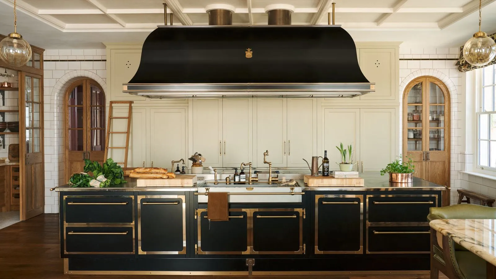 Stort sort og cremet køkken med vintage-indslag. Dens skabe har messingkanter, og den store sorte emhætte over øen er et iøjnefaldende og usædvanligt omdrejningspunkt, der får dette køkken til at føles meget storslået og 1920'er-salonstilen' loading='lazy' title=
