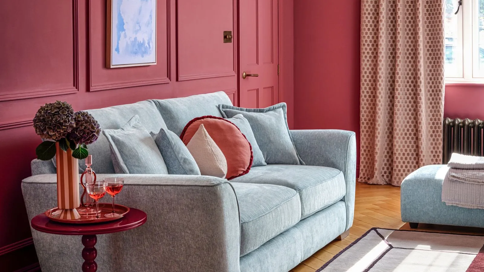 Ein rot bemaltes Wohnzimmer mit hellblauem Sofa mit roten Kissen. Ein kleiner roter Beistelltisch neben der Couch mit einer hohen Blumenvase.' loading='lazy' title=