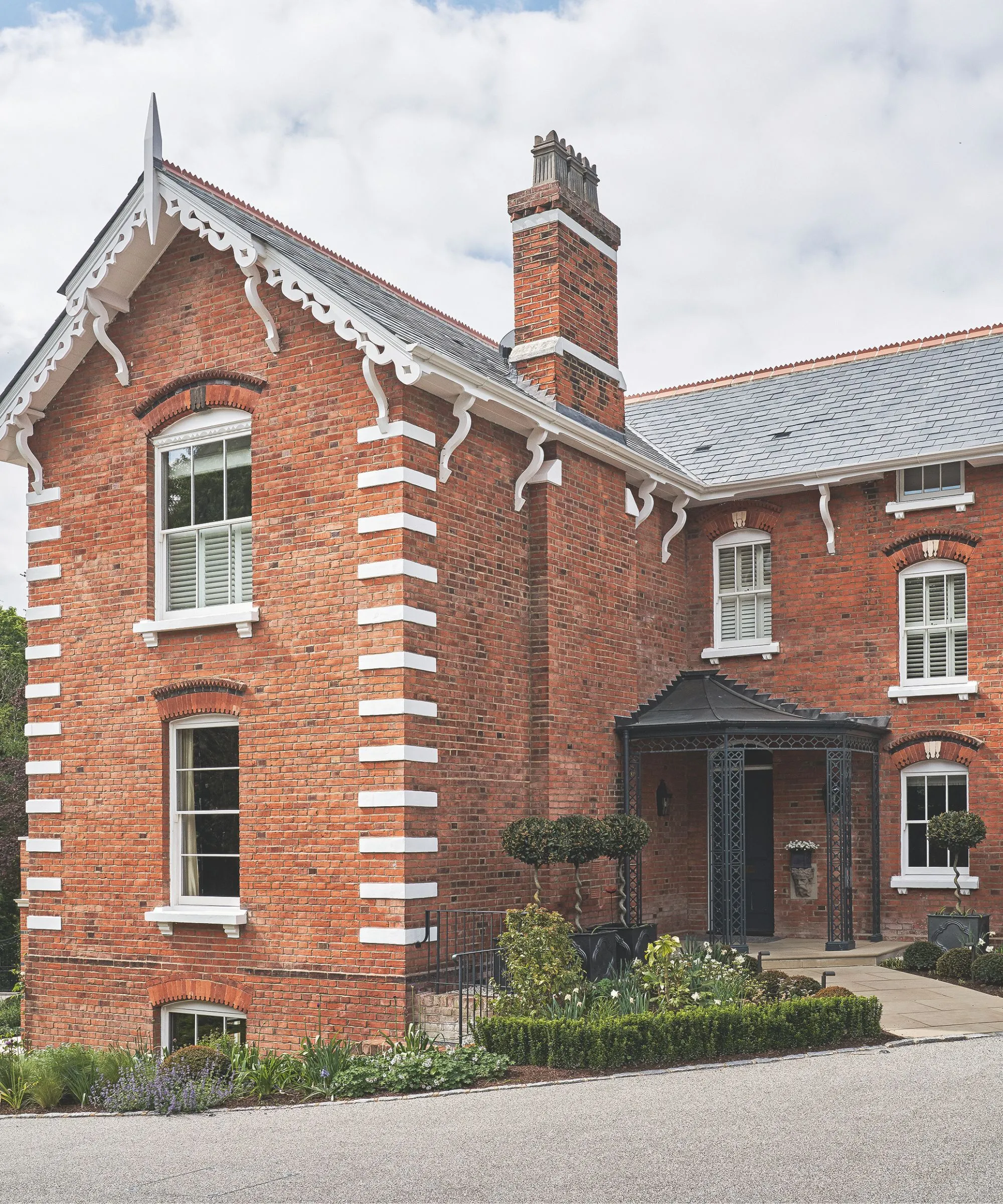 Uma grande casa de tijolos vermelhos com um toldo preto sobre a porta da frente e um jardim frontal bem cuidado com flores e cerca viva baixa.' loading='lazy' title=