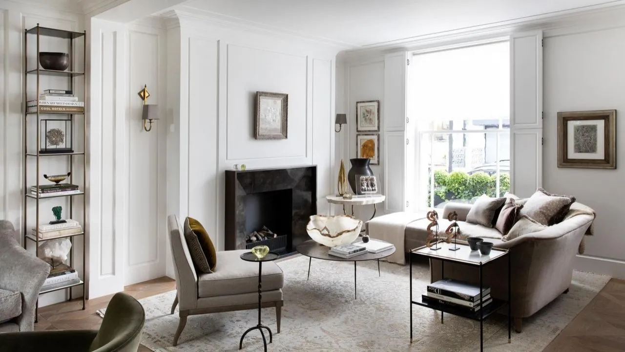 Uma sala de estar branca com móveis neutros, grandes janelas, paredes com painéis brancos, um tapete estampado de estilo persa claro e uma lareira de mármore preto no centro da parede posterior. À esquerda da lareira há uma estante alta com livros e decorações.' loading='lazy' title=