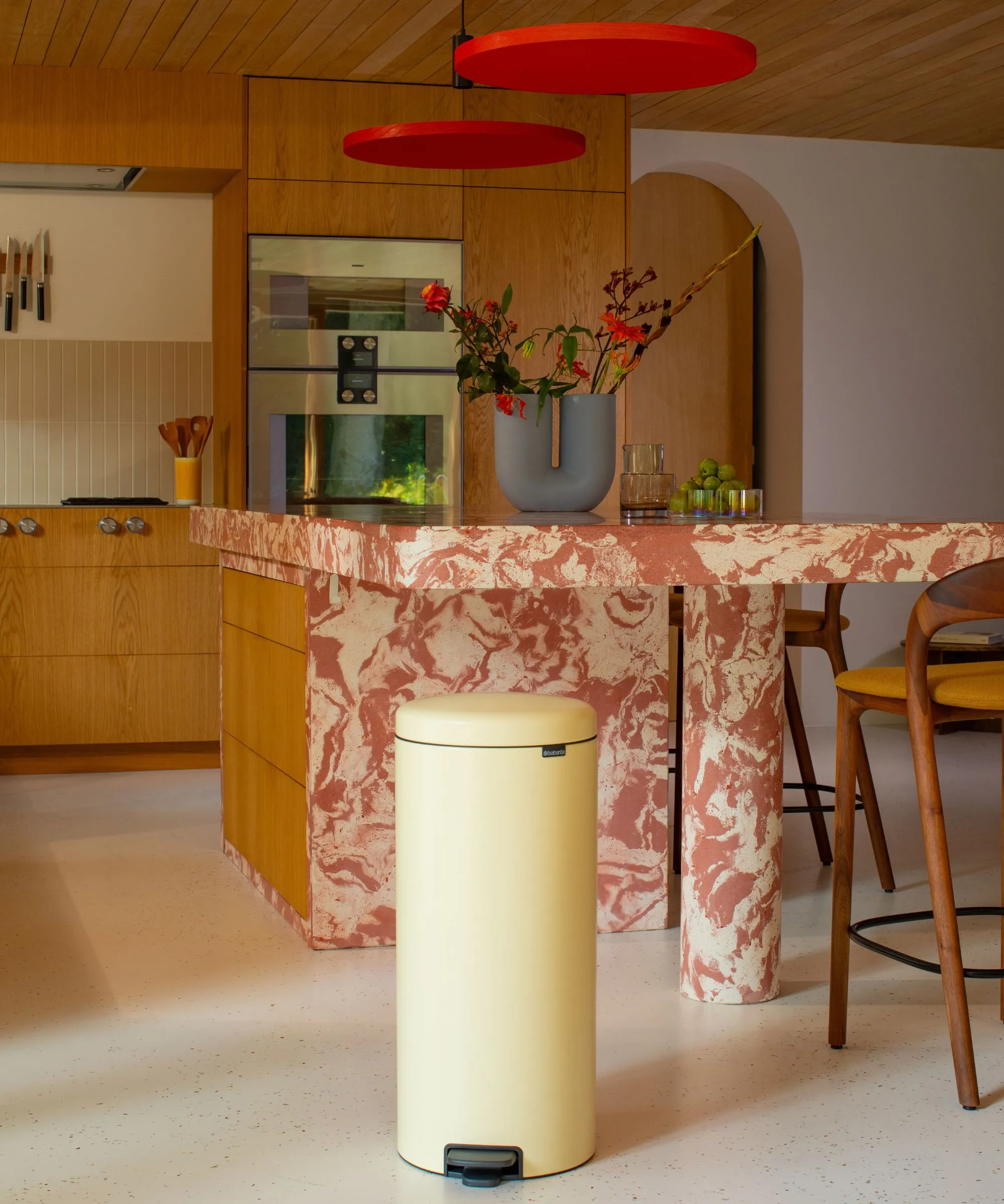 Kuchnia z marmuru i drewna z maślanożółtym koszem na śmieci na pierwszym planie blatami z czerwonego marmuru i drewnianymi szafkami.' loading='lazy' title=