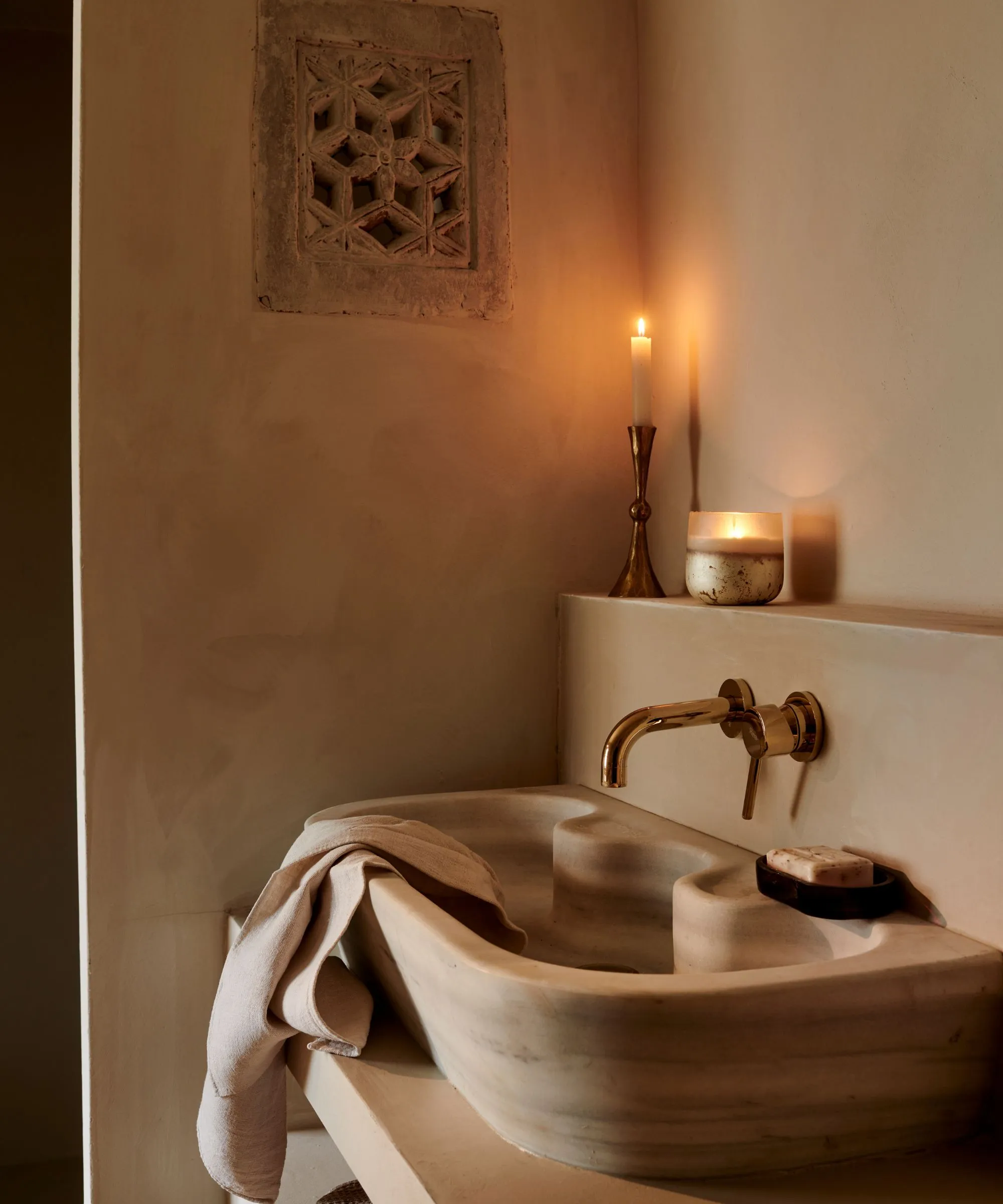 En udskåret stenvask i et badeværelse med pastervæg. Et lys tændt på afsatsen ovenfor.' loading='lazy' title=